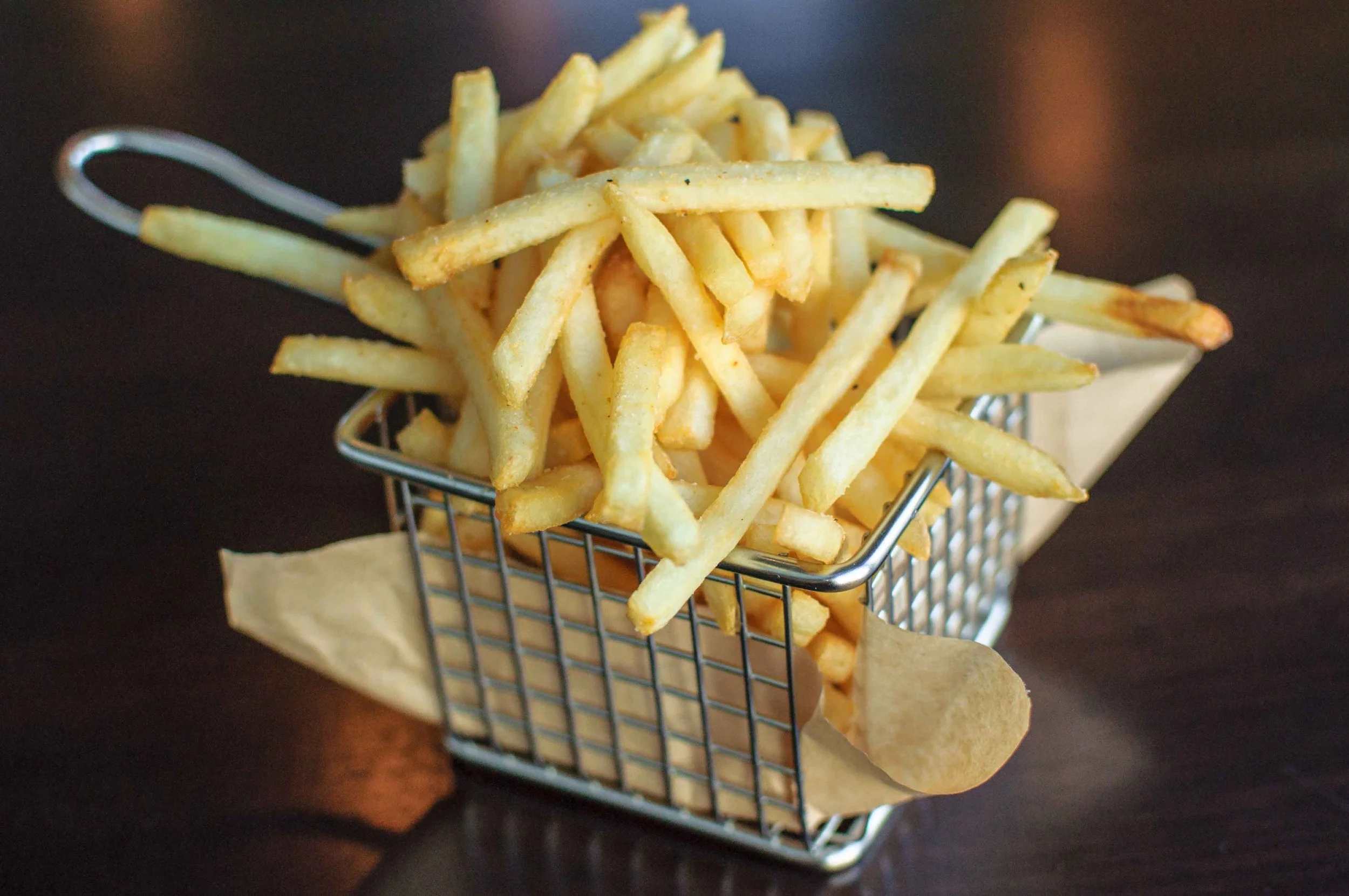 French fries in a small metal basket lined with paper on a dark surface.