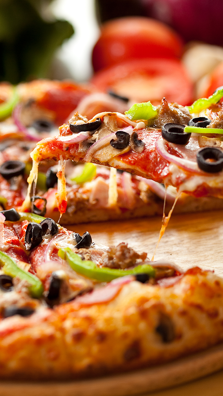 Close-up of a slice of pizza with black olives, green peppers, onions, sausage, and melted cheese, on a wooden surface with whole tomatoes and other produce in the background.