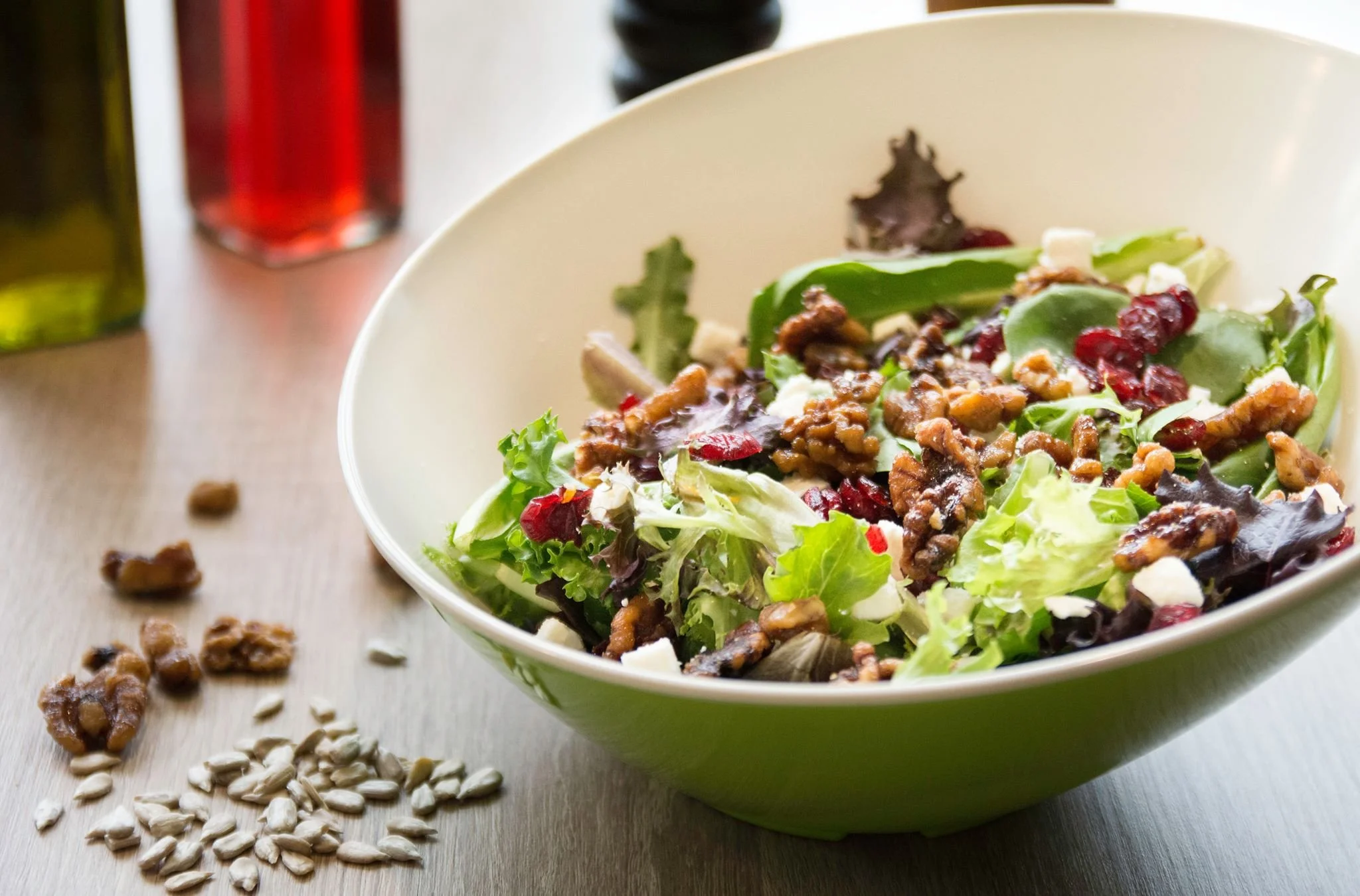 A bowl of salad with greens, nuts, dried cranberries, and cheese, on a wooden surface with sunflower seeds and walnuts spilled around.