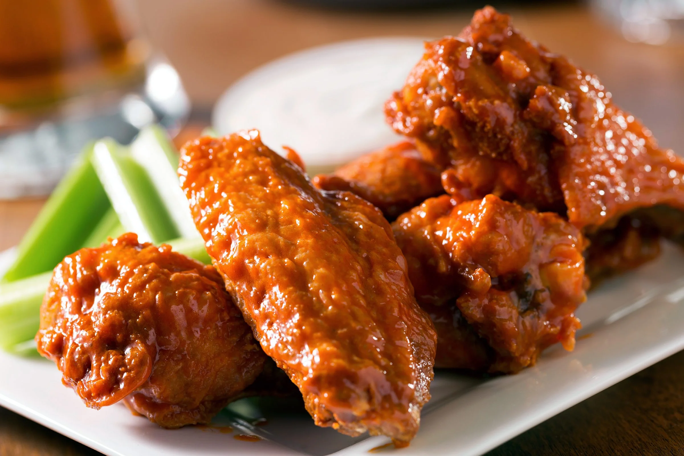 Close-up of spicy buffalo chicken wings coated in hot sauce on a white rectangular plate, with celery sticks and a blue cheese dressing in the background.