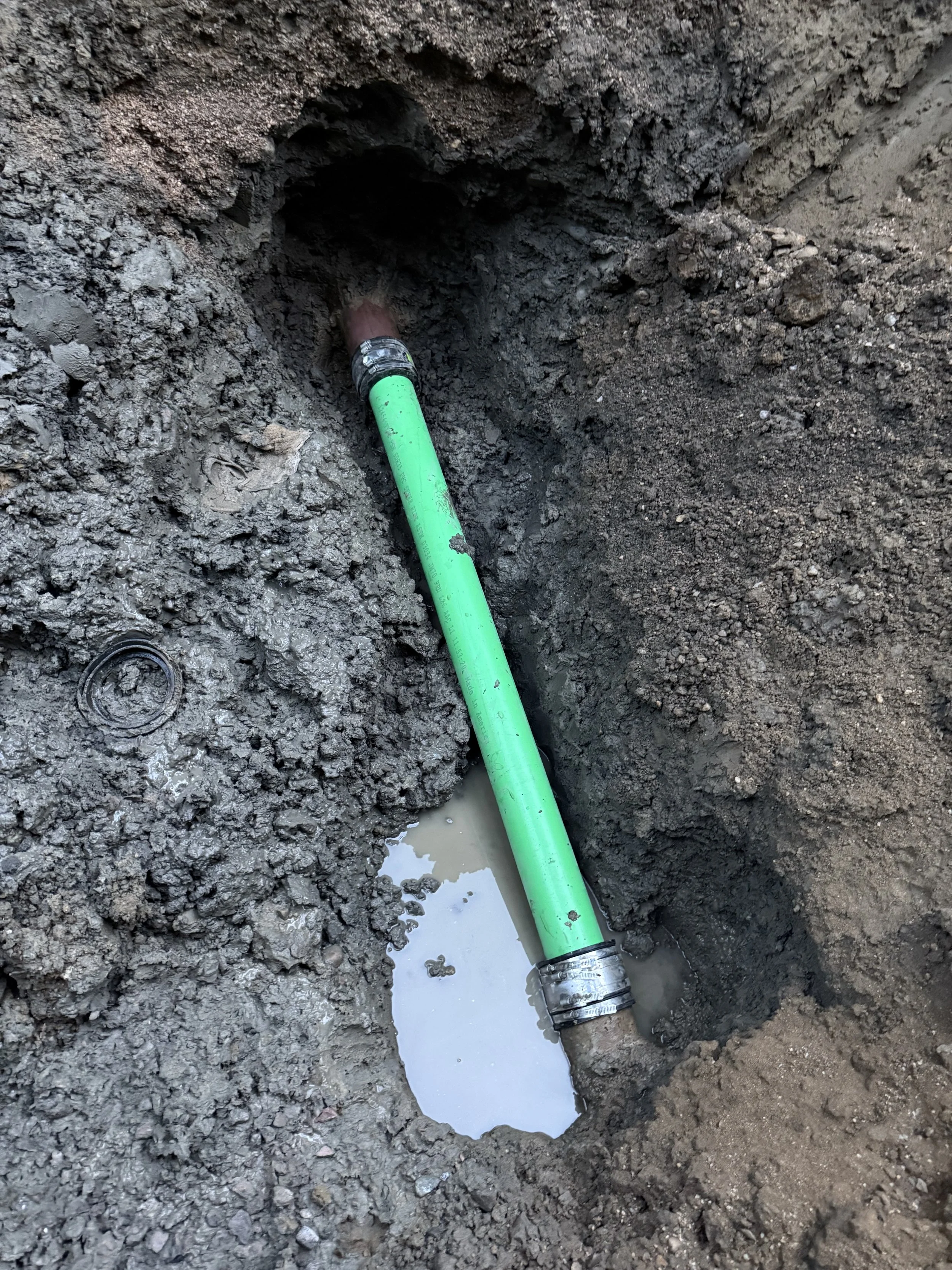 A green pipe connected to a sewer line in a trench filled with dirt and water.