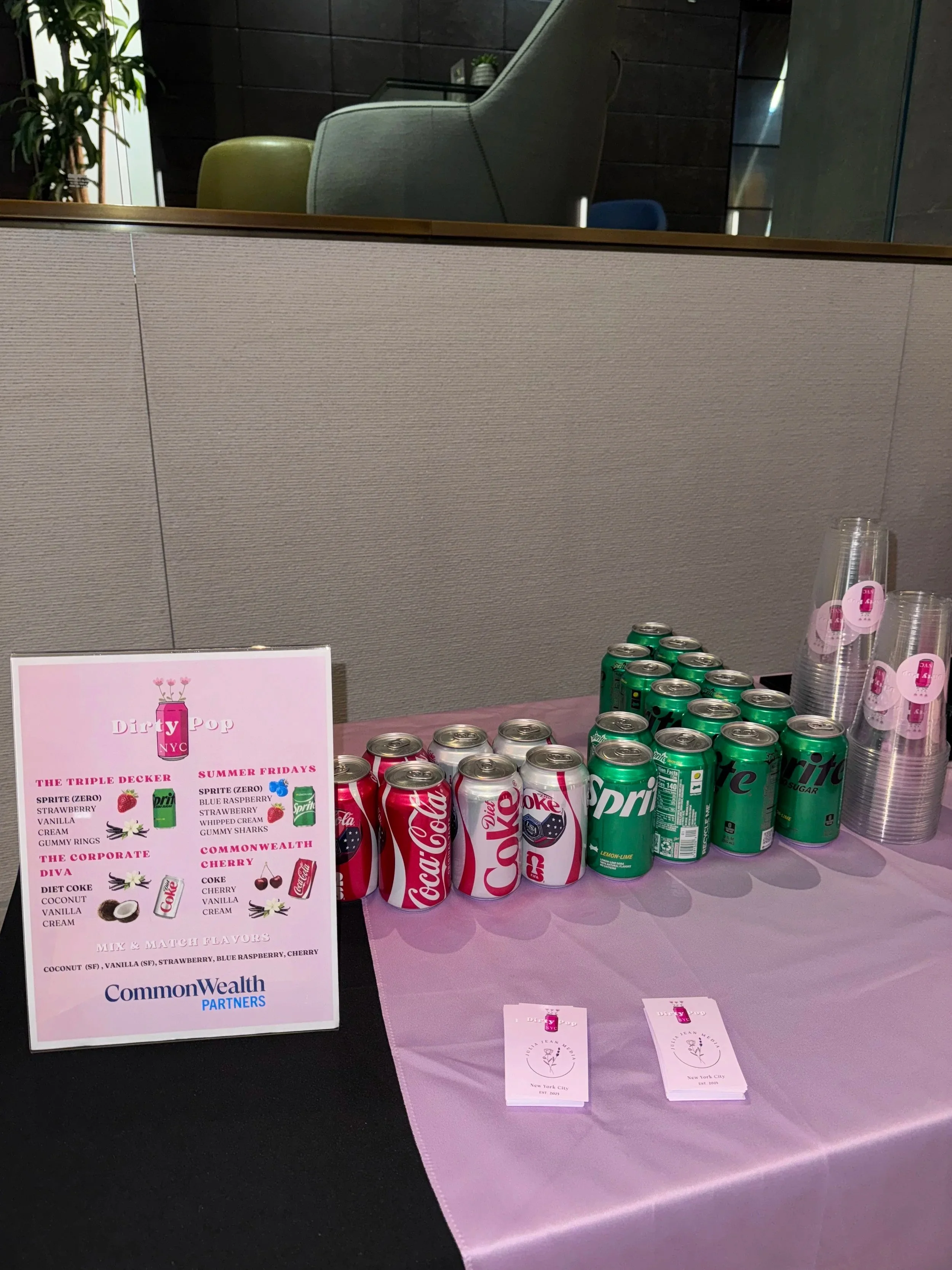 Table with various soda cans, plastic cups with pink labels, and a printed menu, in a modern indoor setting with chairs and a plant in the background.