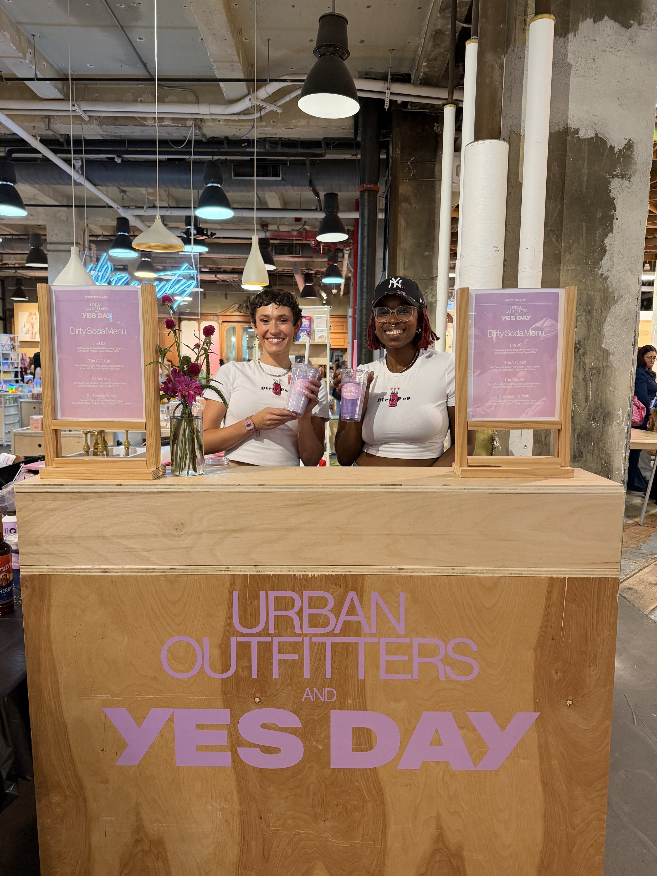Two women standing behind a wooden counter at a store event, smiling and holding cups with straws. There are purple signs on each side of the women, and large pink letters on the front of the counter that read 'URBAN OUTFITTERS' and 'YES DAY.'
