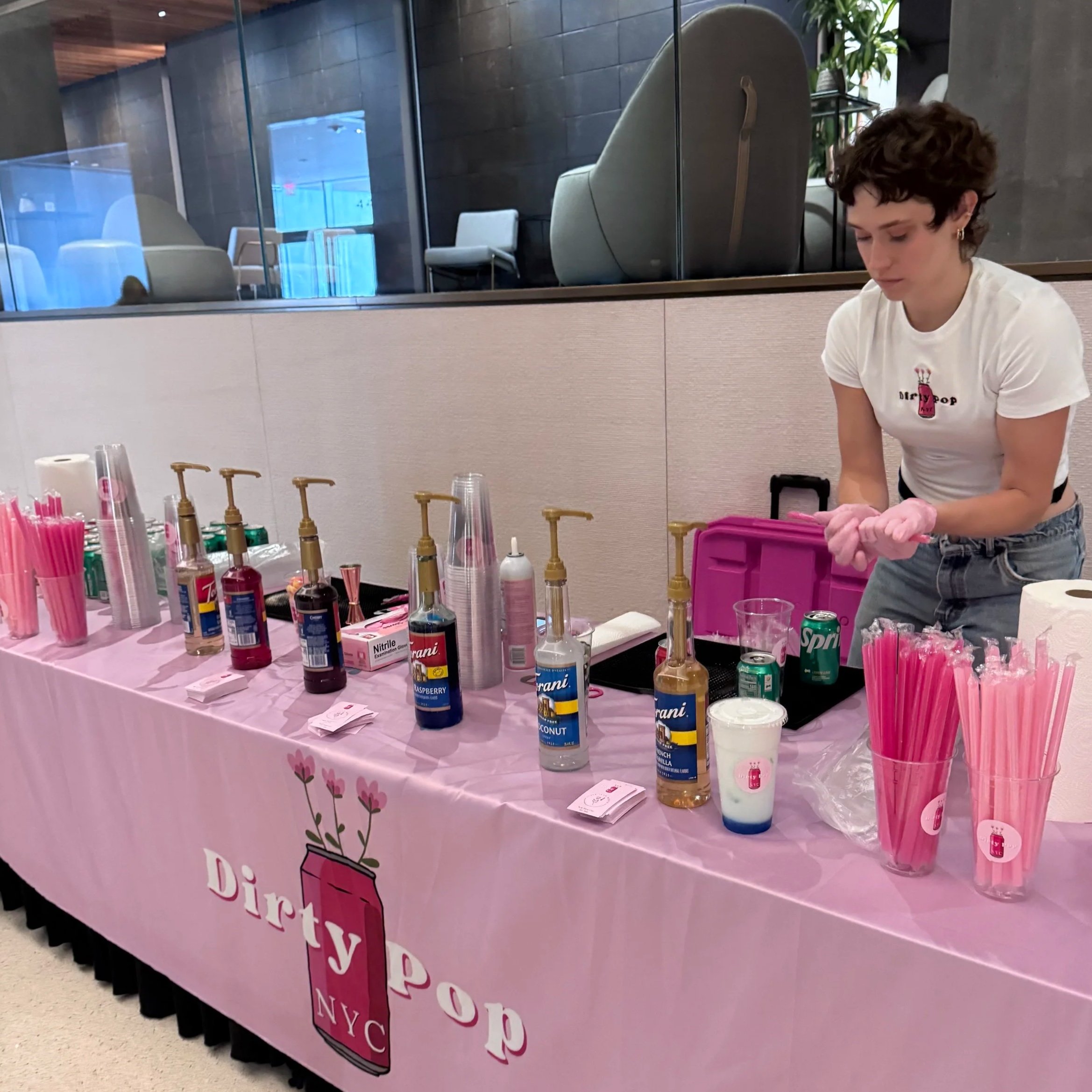 A pink tablecloth with a floral design covers a table displaying bottles of nail polish, brushes, and other nail care supplies at a beauty event. Behind the table, there is a glass partition dividing the space, with a woman in a white T-shirt preparing items.