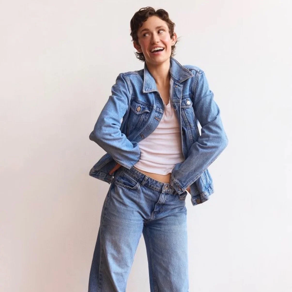 A young woman wearing a denim jacket and jeans, smiling and standing against a plain white background.