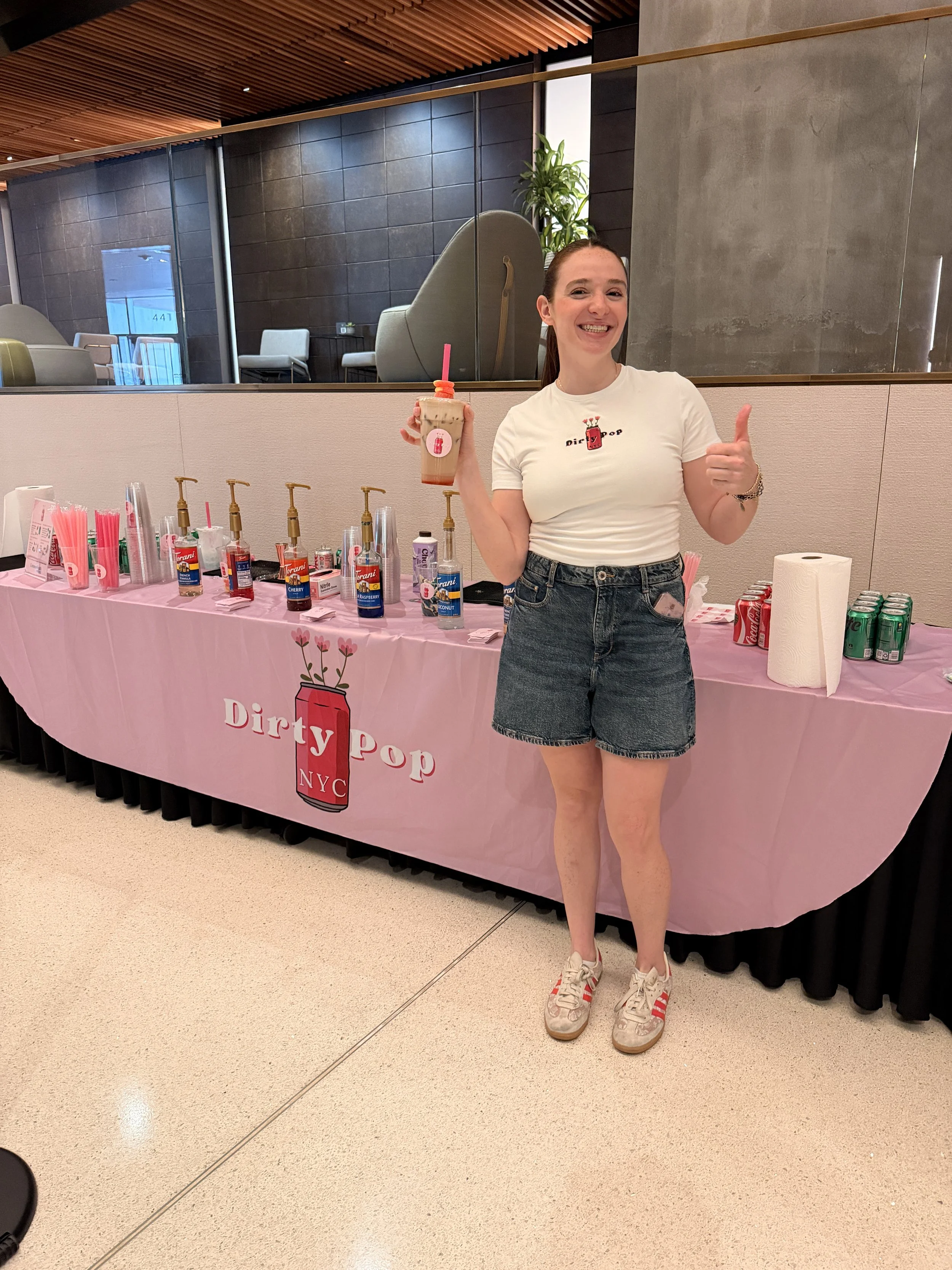A young woman with brown hair in a ponytail, wearing a white t-shirt, denim shorts, and sneakers, standing in front of a pink table with various drink bottles and cups, holding a pink drink with a straw and giving a thumbs-up gesture, in a modern indoor setting.