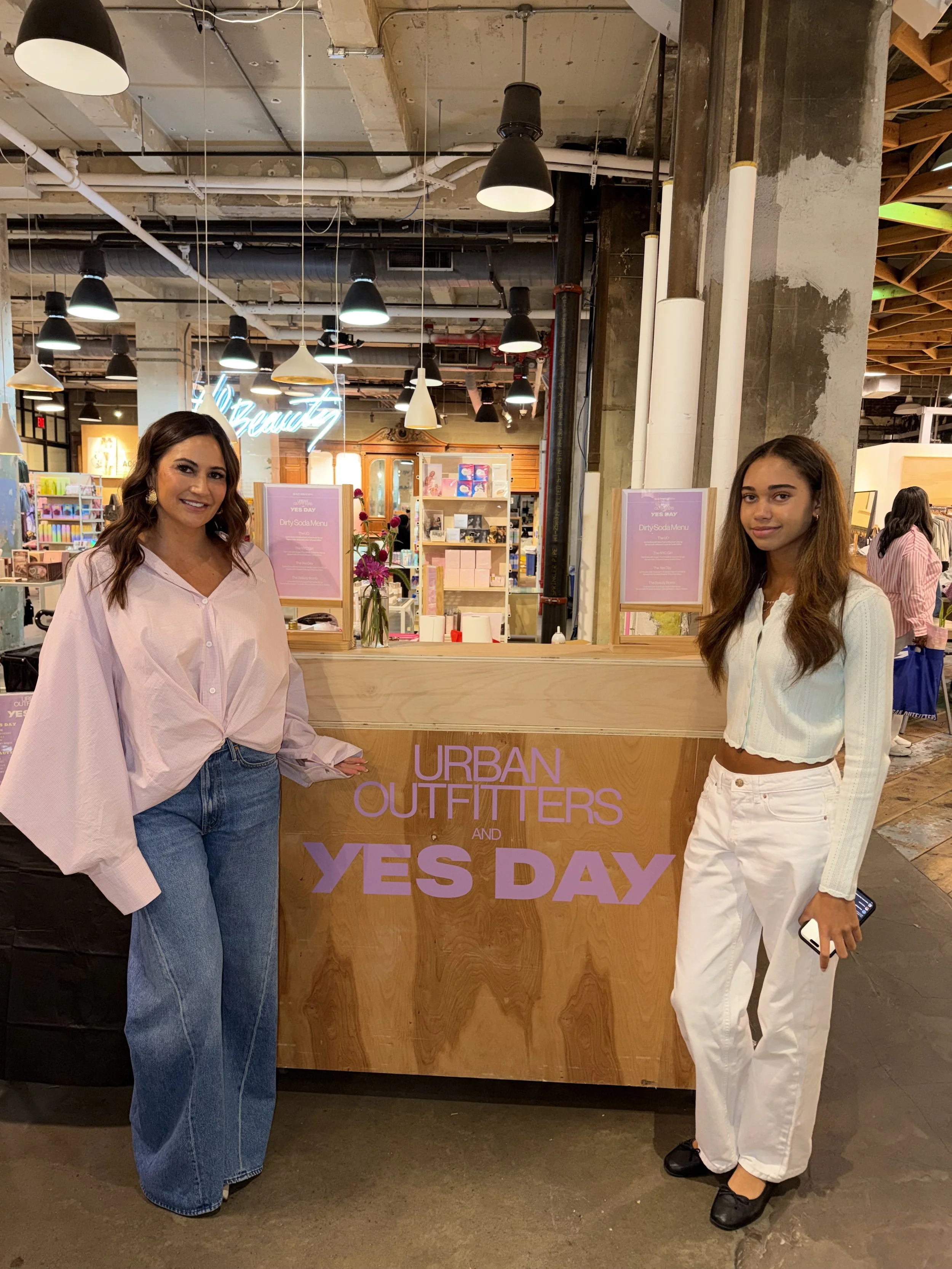 Two women standing in front of a wooden counter with a sign that reads 'Urban Outitters and Yes Day,' inside a retail store with industrial ceiling and hanging lights, smiling at the camera.