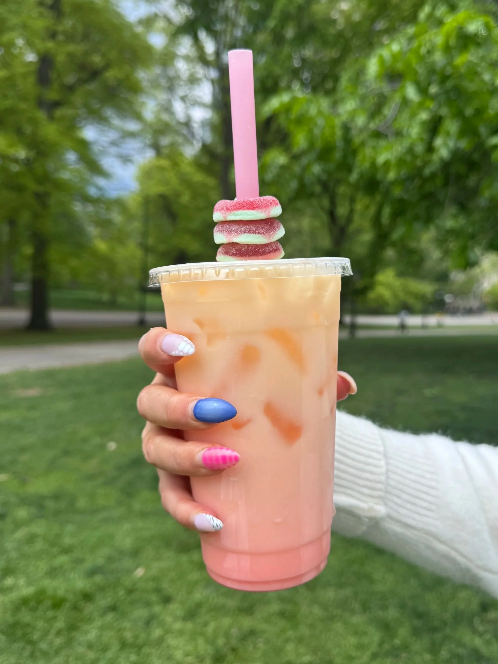 A person holding a peach-colored drink with whipped cream and apple slices on top, with a pink straw, in an outdoor park setting.