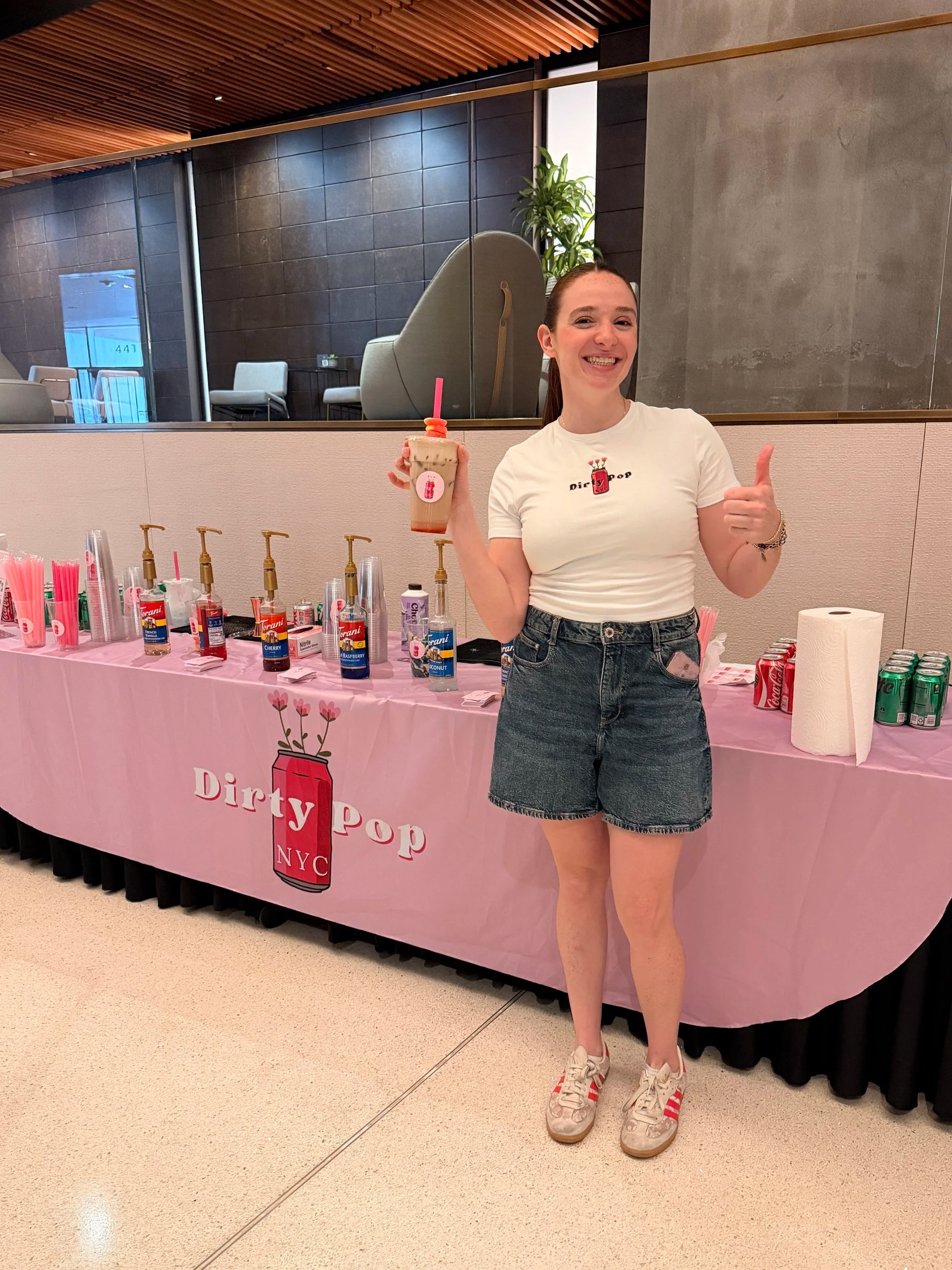 A woman smiling and giving a thumbs-up while holding a drink at a pink booth with the text 'Dirty Pop NYC' on it. The booth has various syrups and cups, with paper towels and cans of soda on the side.