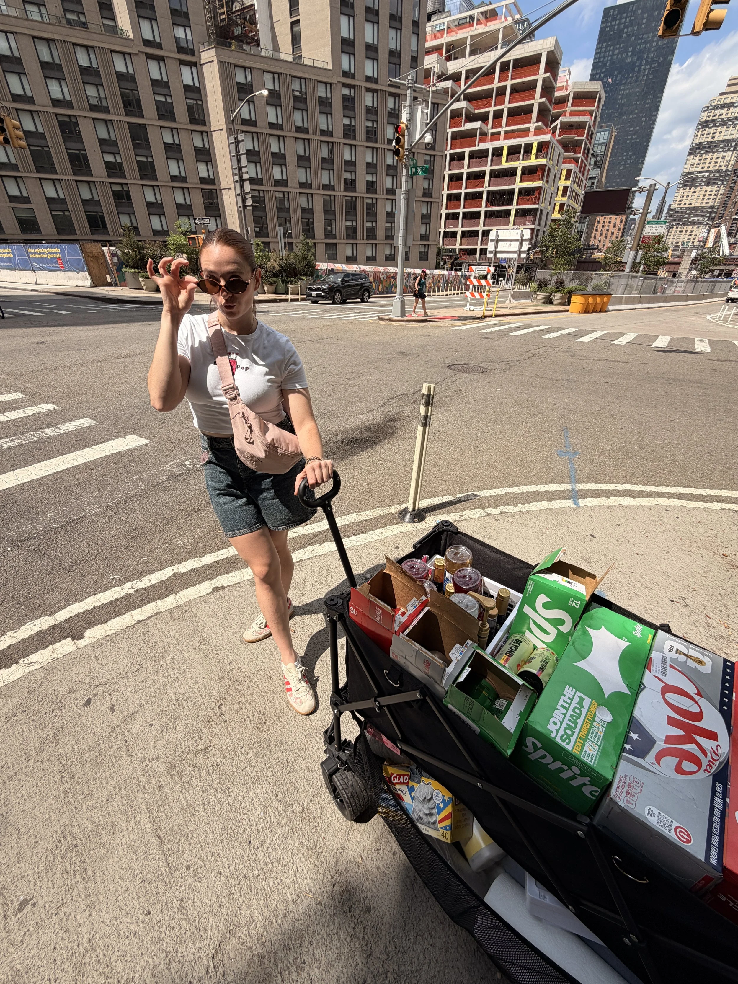 Woman wearing sunglasses and casual clothing, pushing a shopping cart filled with grocery items on a city street with tall buildings, construction site, and crosswalks in the background.