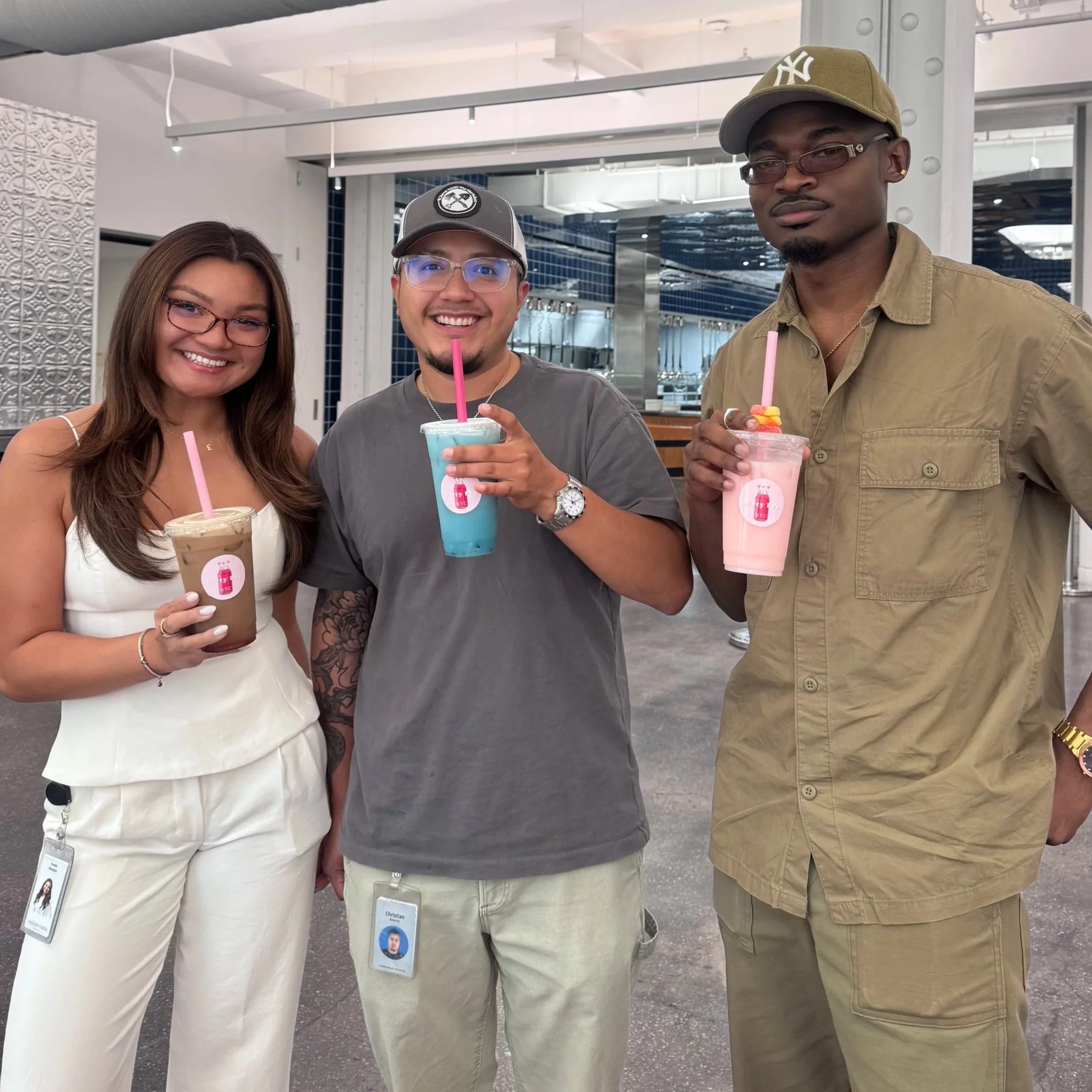 Three young adults smiling and holding colorful drinks in a modern indoor setting with glass and metal decor.