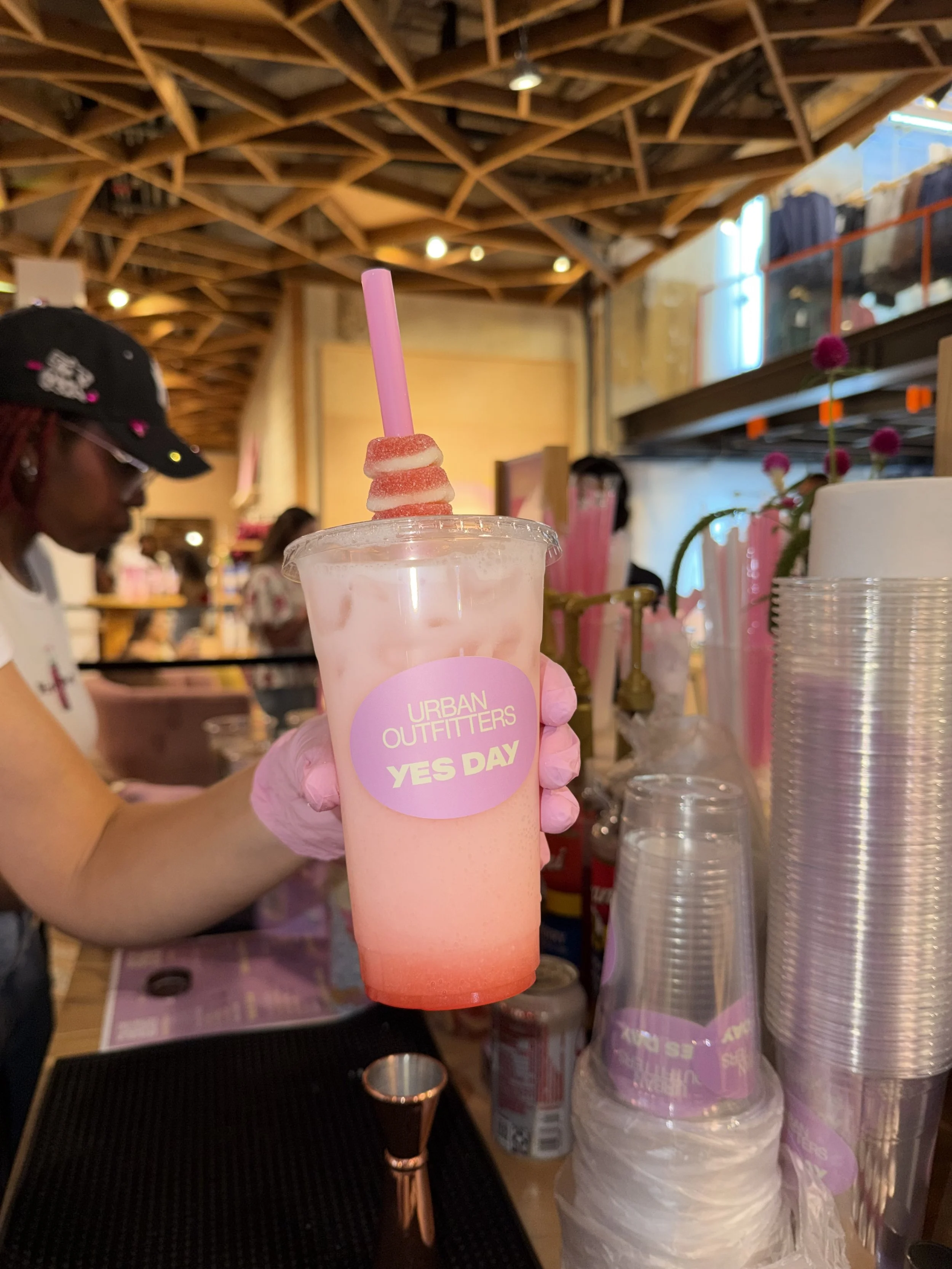A person in a pink glove holding a pink and white layered beverage with a pink straw at a gathering or event, with other people and supplies visible in the background, and a wooden ceiling above.