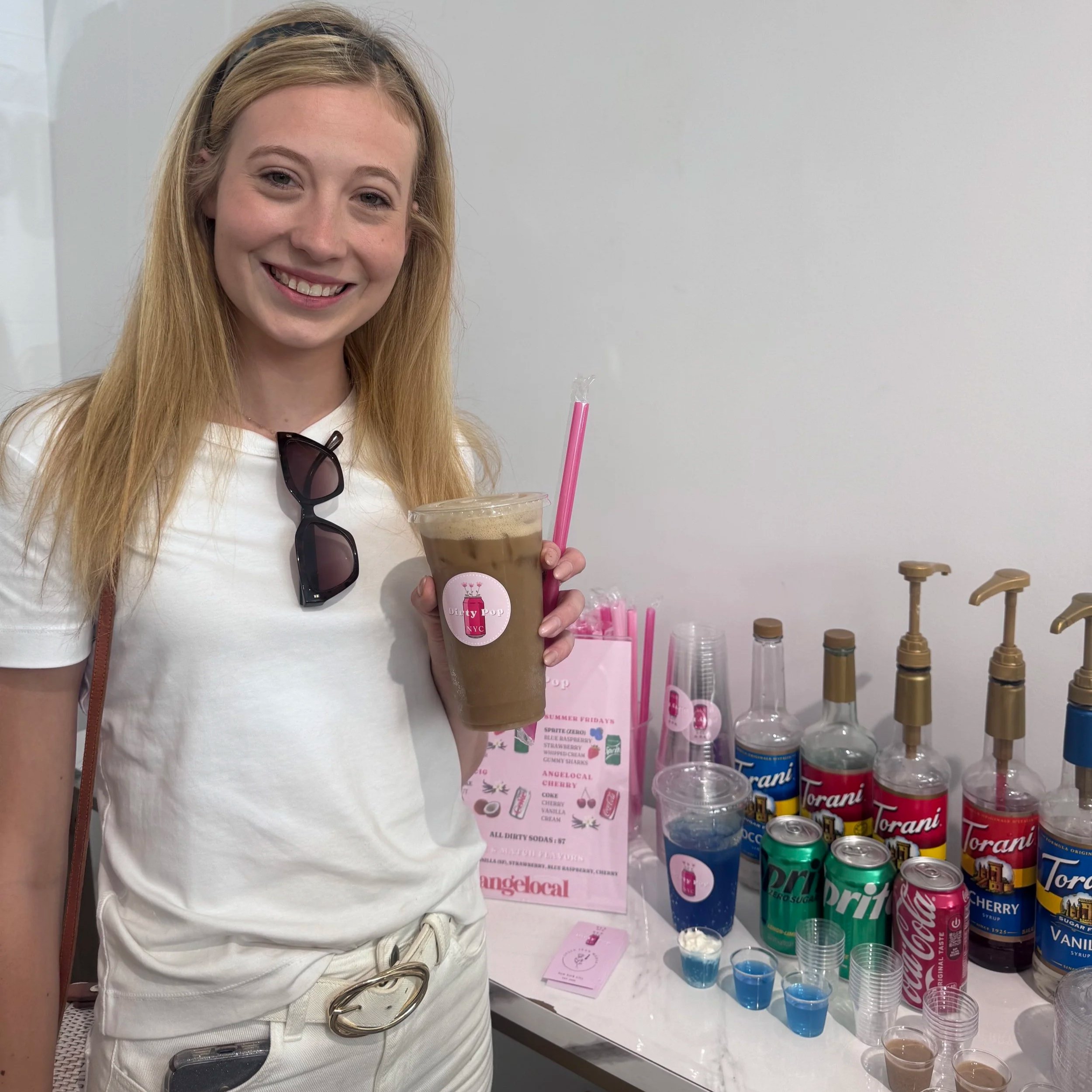 A young woman with long blonde hair and a headband smiling while holding a clear plastic cup filled with a layered chocolate and vanilla drink, standing in front of a white wall and a table with various drink ingredients and colorful soda cans.