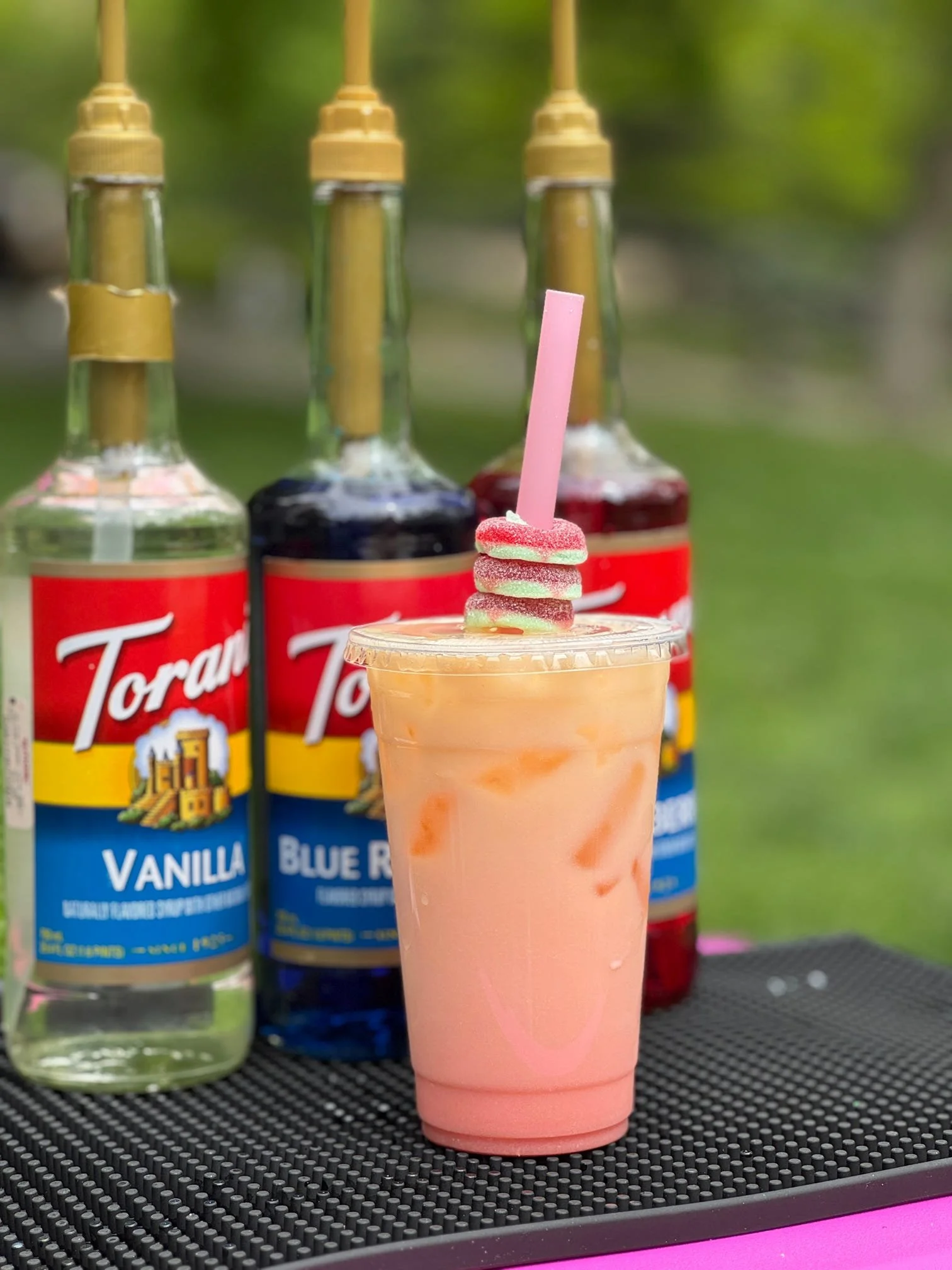 A pink smoothie topped with three colorful gummy candies and a pink straw, with bottles of Torani vanilla and blue raspberry syrups in the background.