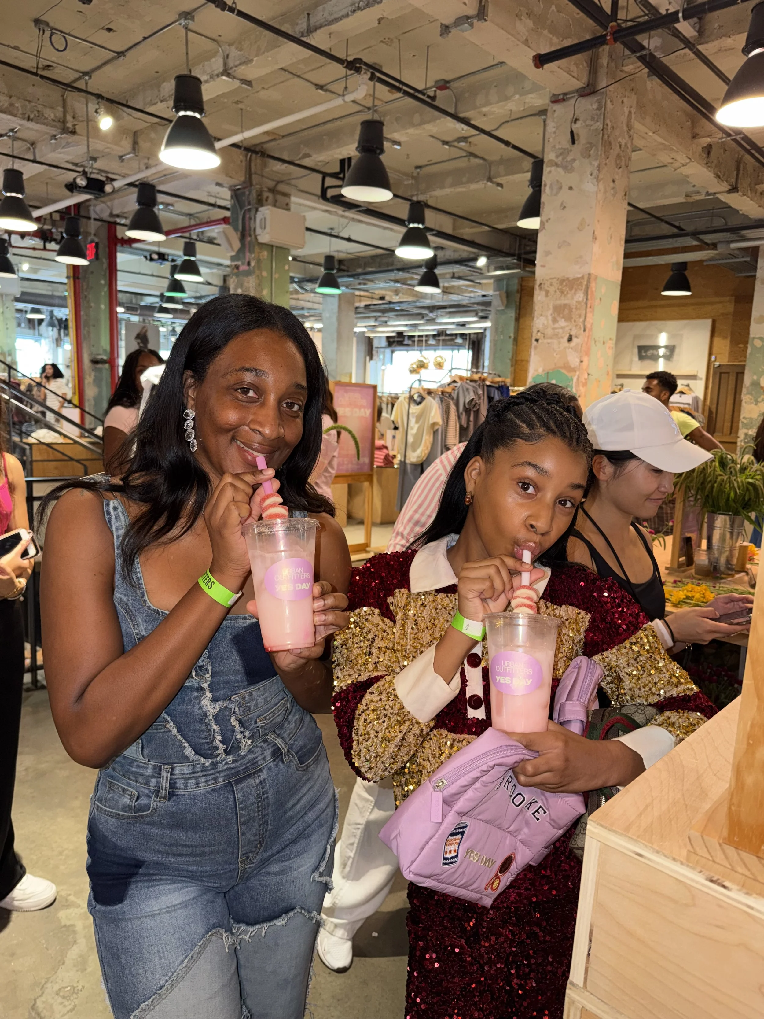 Two women holding pink drinks with straws, standing in a busy indoor market or mall, smiling and enjoying their beverages. One woman is wearing a denim dress, and the other a sparkly red dress with an aqua pink backpack.