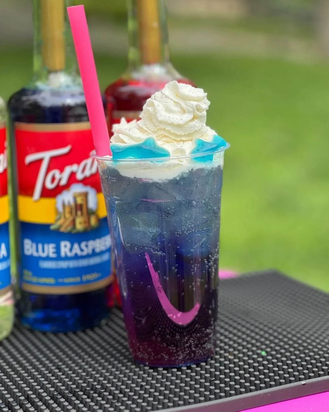 A tall glass of blue raspberry soda with whipped cream, blue gummy candies, and a pink straw, placed on a black textured surface with bottles of Torani blue raspberry syrup in the background.
