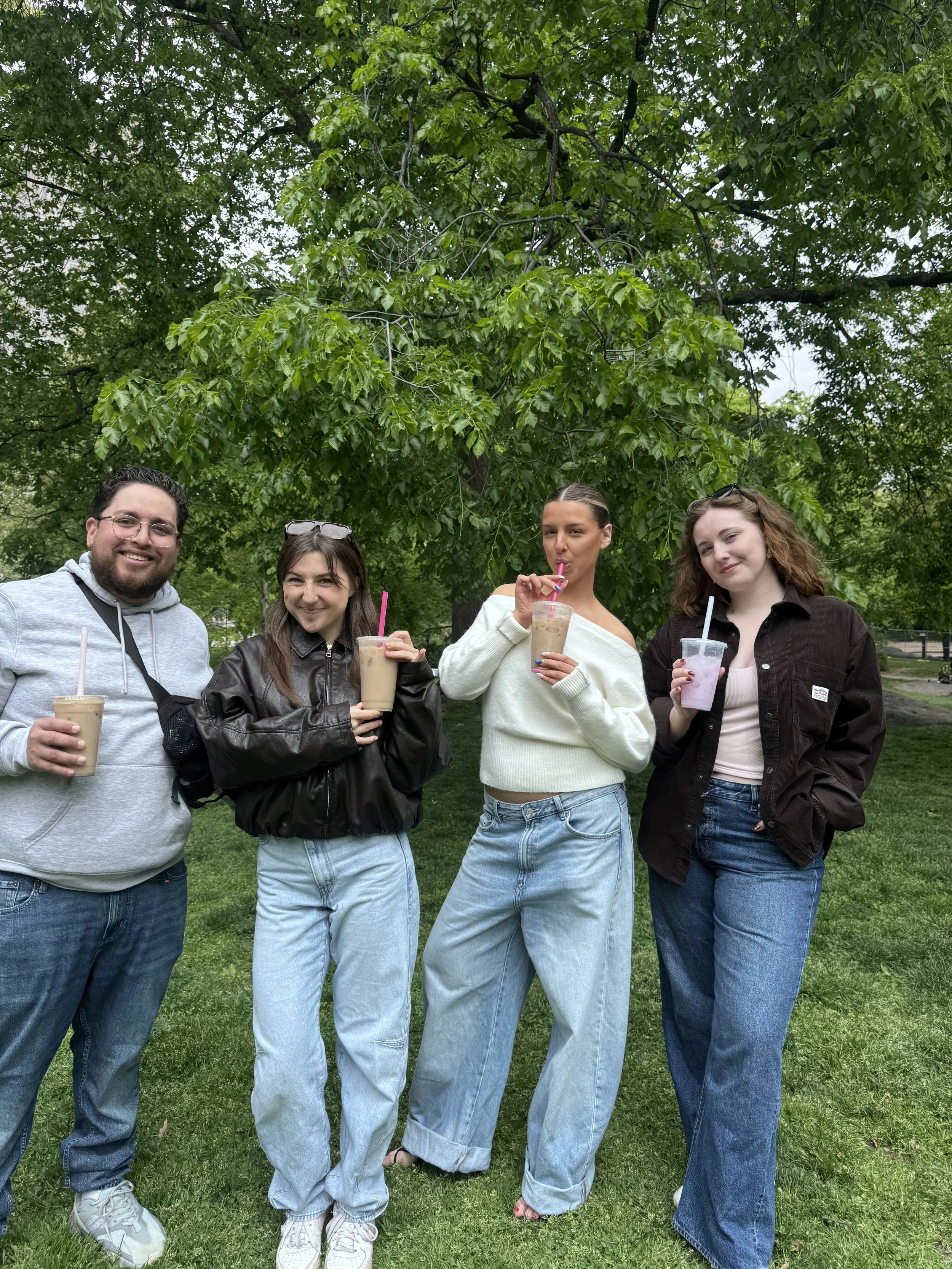 Four young adults standing outdoors on grass in front of a large leafy green tree, holding various beverages and smiling.