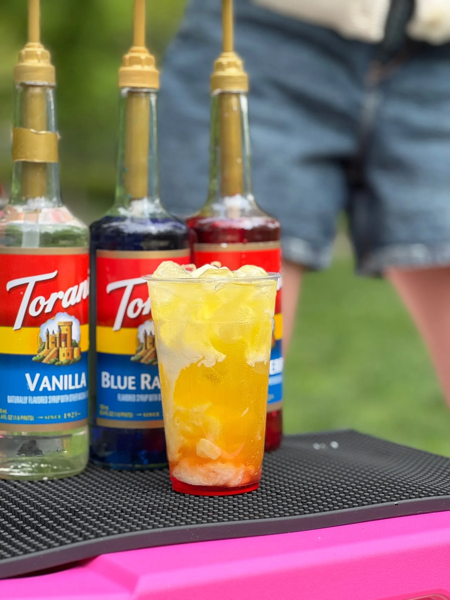 A colorful drink with ice and slices of fruit in a plastic cup in the foreground. Behind it, there are four bottles of flavored syrup labeled vanilla and blue raspberry, with yellow lids, on a black textured surface outdoors.