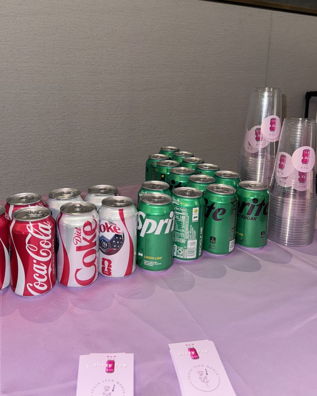 Soft drinks, including Coca-Cola, Diet Coke, Sprite, and Lime Sprite, arranged on a pink table with clear plastic cups stacked with pink and white stickers.