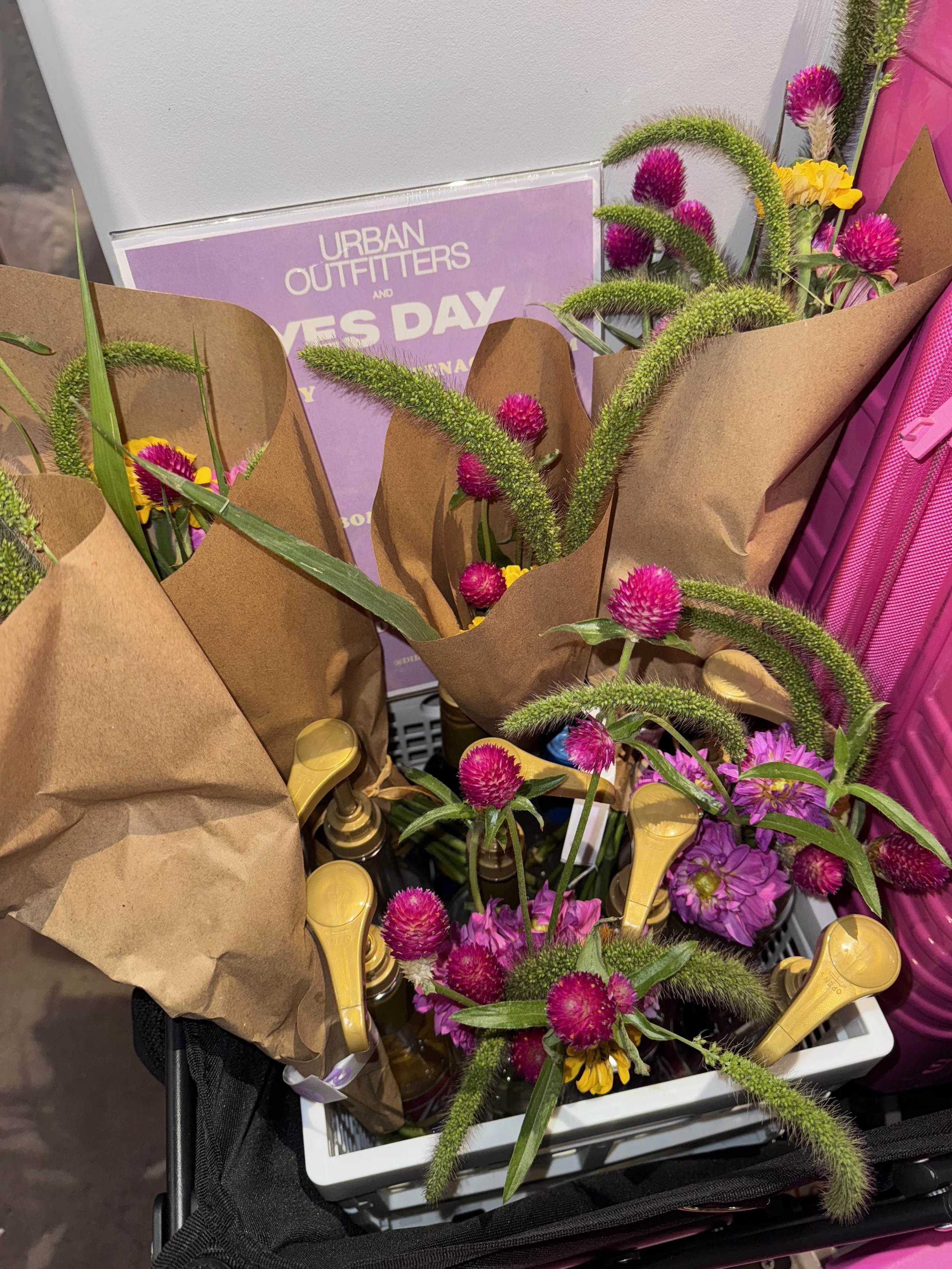 A shopping cart filled with bouquets of colorful flowers, including pink globe amaranth and yellow and purple flowers, with greeting cards and bottles of wine or champagne.