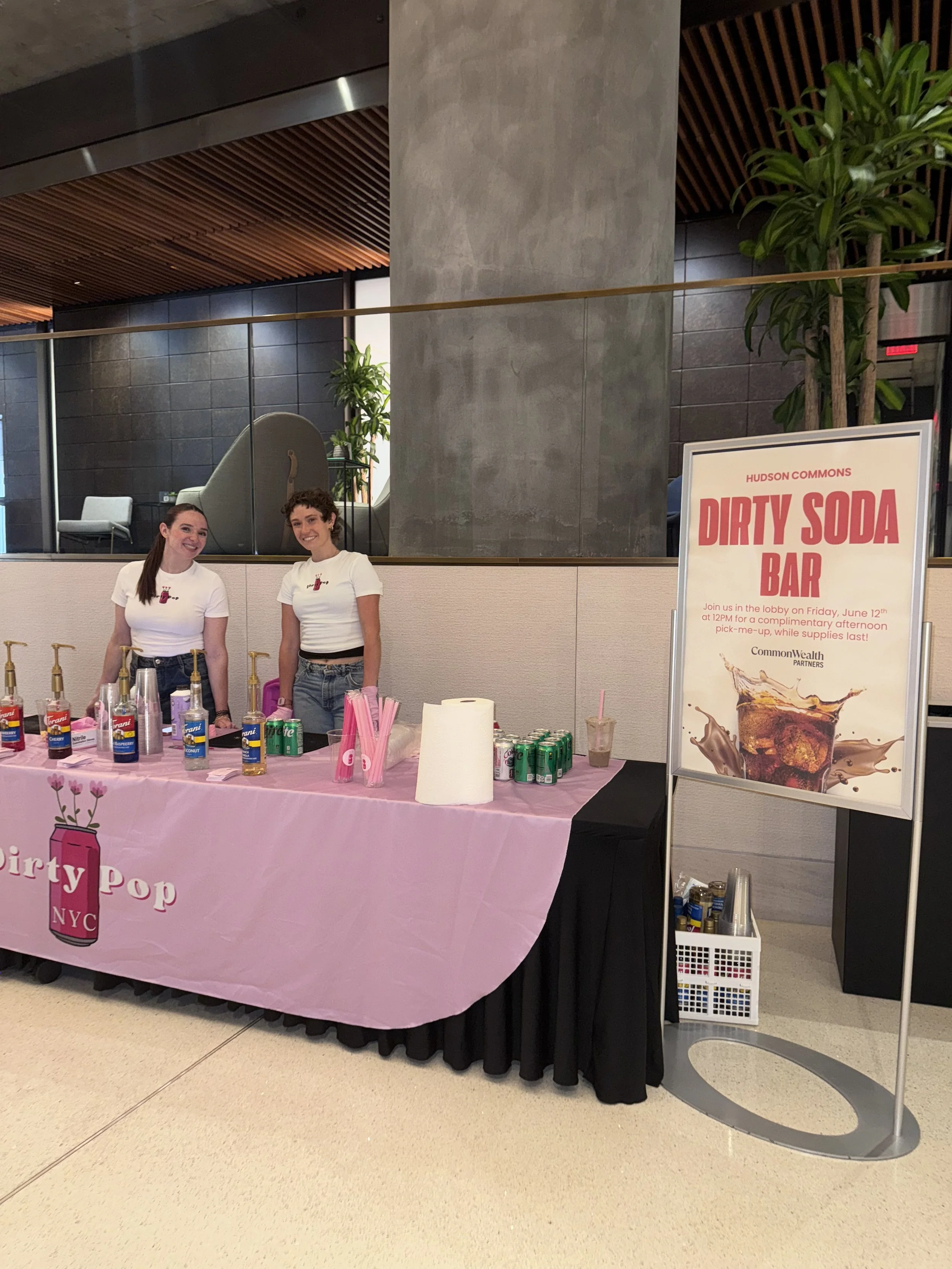 A pink table with a logo that says "Dirty Pop NYC" and pictures of pink soda cans and cups with pink straws. Two women with long hair, wearing white T-shirts and jeans, standing behind the table. A sign next to them promotes a "Dirty Soda Bar" event at Hudson Commons.