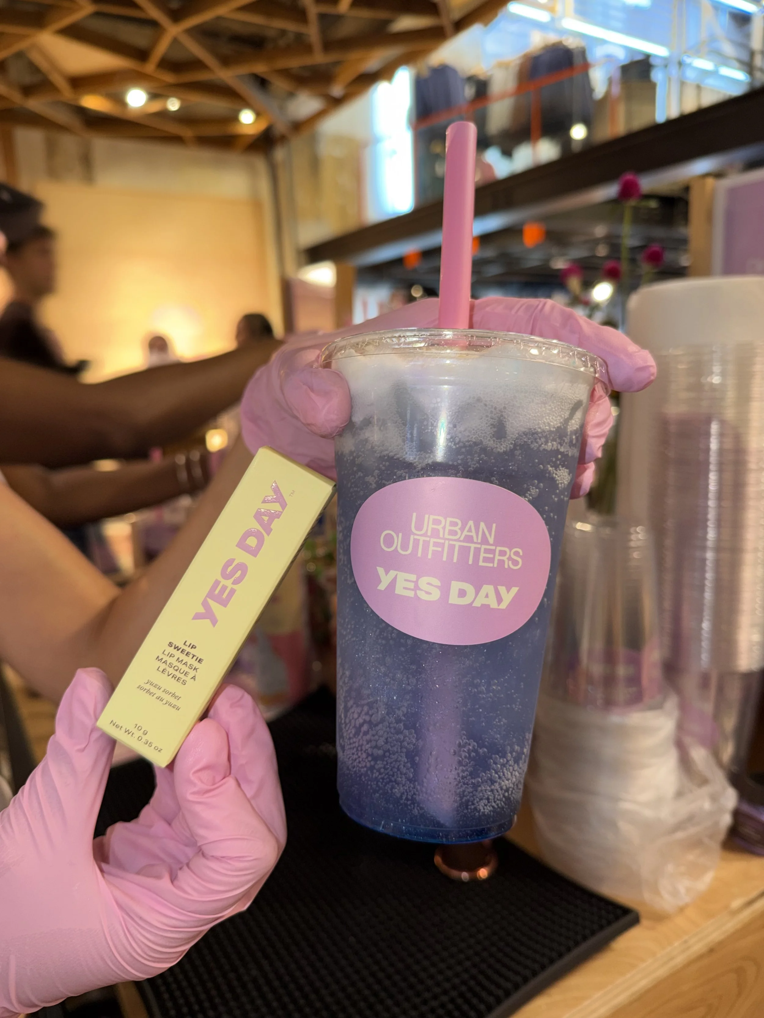 A person wearing pink gloves holds a clear plastic cup filled with a dark purple beverage, with a pink straw. The cup has a pink sticker reading 'Urban Outfitters Yes Day.' The person also holds a yellow tag. The background reveals a wooden ceiling and blurred store or cafe environment with other people and products.