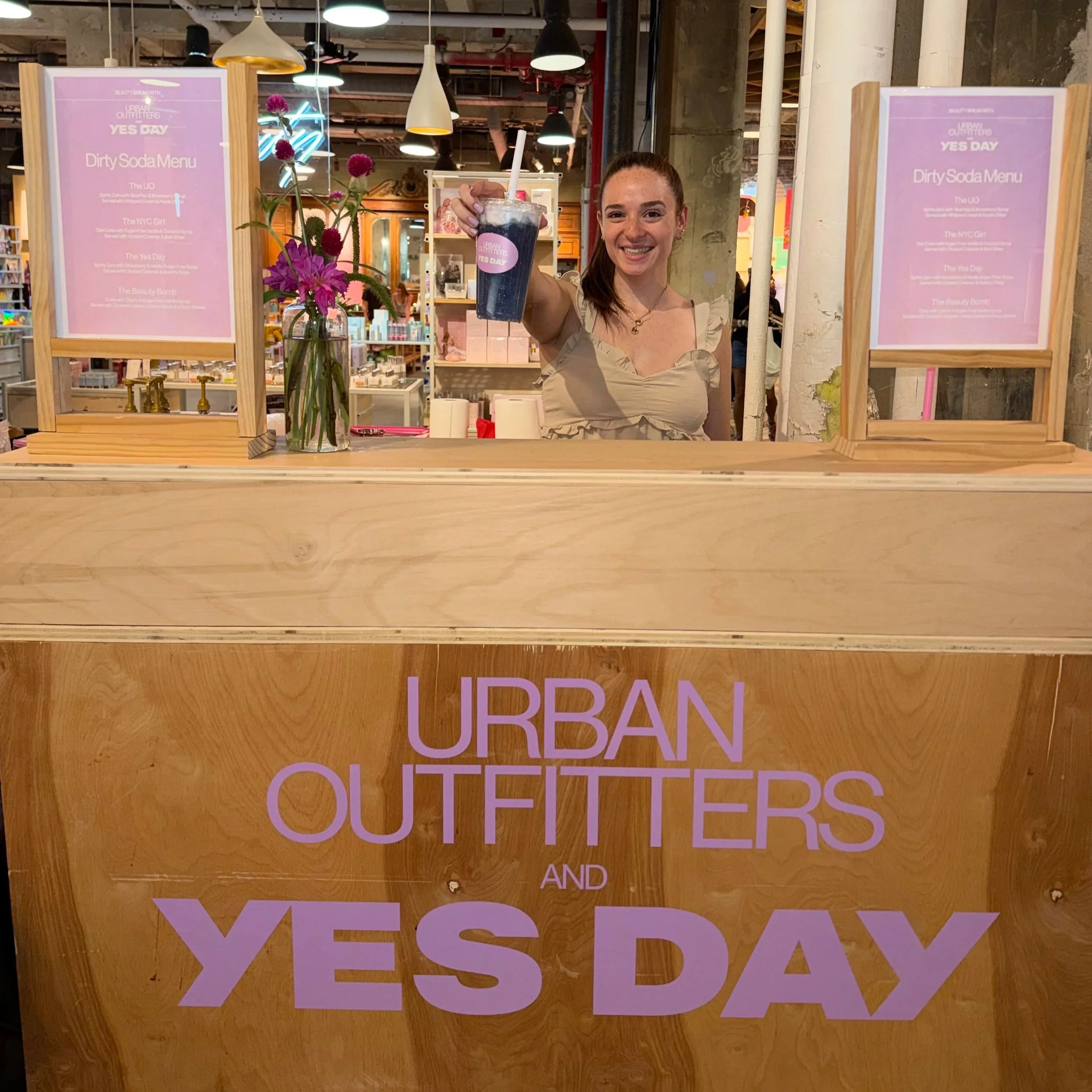 A woman smiling and holding a dark soda in a clear cup with a straw at an indoor event with pink signs and wooden decor.