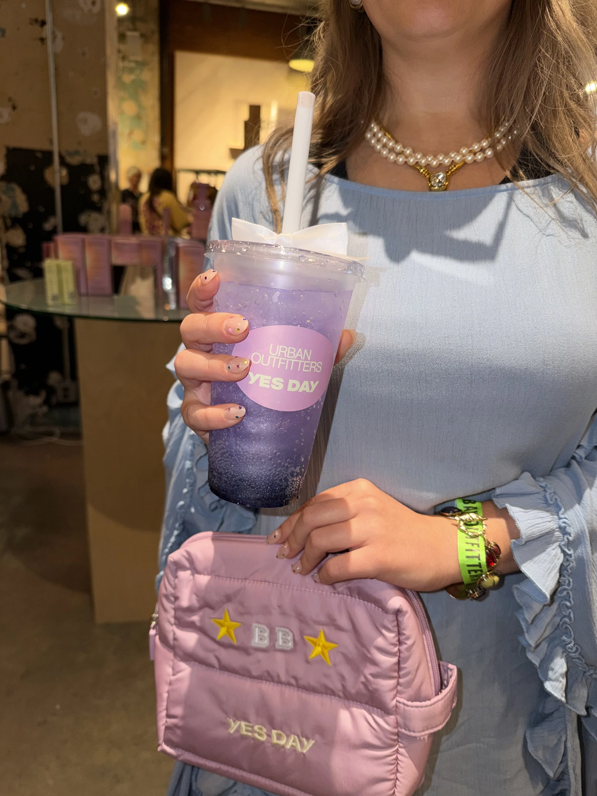 A woman holding a purple, star-decorated drink cup with a straw, wearing a pearl necklace and a light blue top. She has a pink quilted purse with yellow stars and the words "YES DAY" on it, and a yellow wristband. A bracelet with charms is on her right wrist, and her nails are painted with multicolored dots.