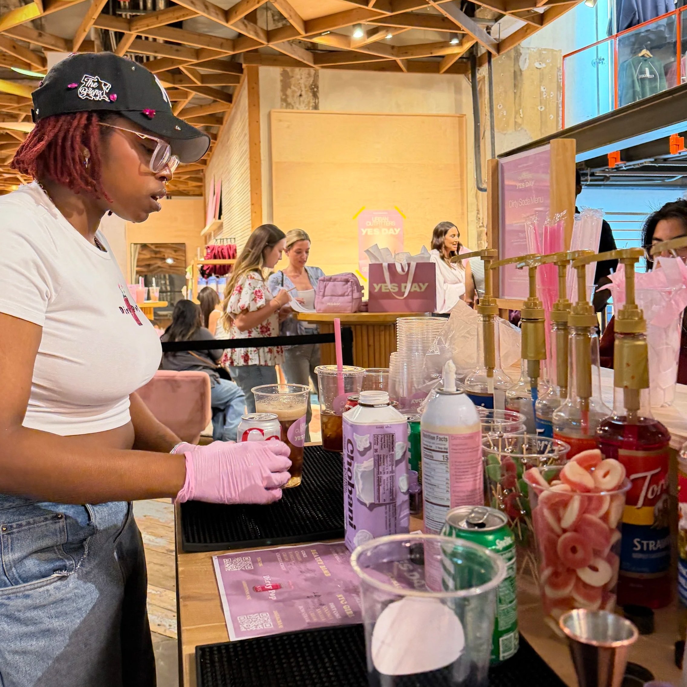 A woman with red dreadlocks, wearing glasses, a black cap that says 'The Drag' and white t-shirt, stands at a counter with various drink ingredients and snacks in a bustling indoor store with wooden ceiling beams, other customers, and promotional posters.