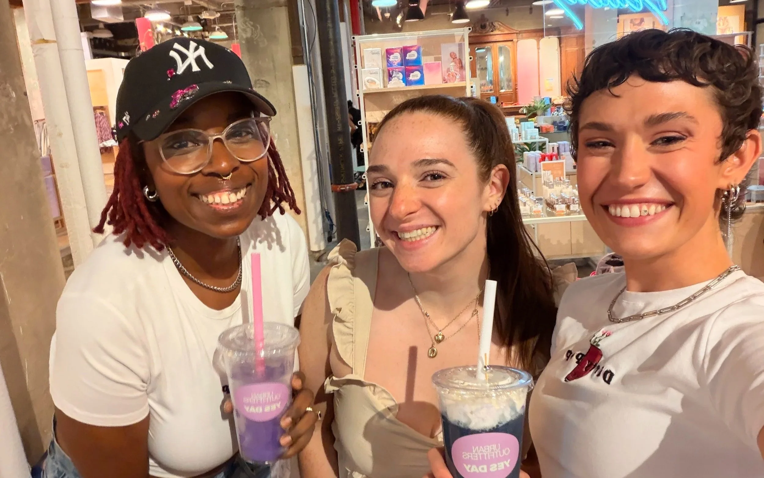 Three women smiling and posing for a photo in a store or cafe, holding drinks with pink straws. The background includes shelves with various products and a wooden cabinet.