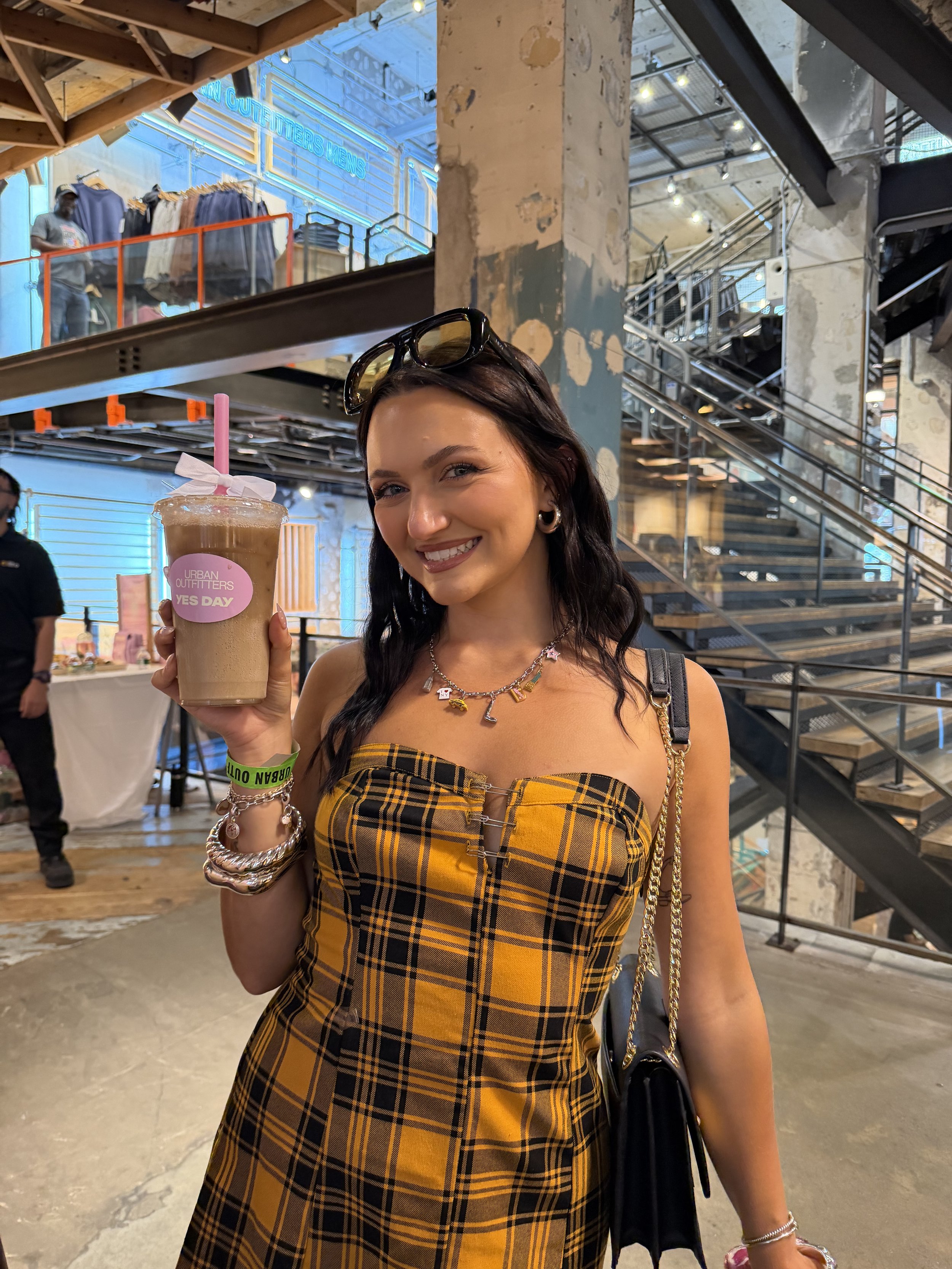 A young woman with dark hair and light skin smiling, holding a large iced coffee in a plastic cup with a pink straw and a paper sleeve. She is wearing a yellow and black plaid dress, several bracelets, a necklace, and sunglasses on her head. The background is a multi-level indoor space with exposed industrial decor, metal stairs, a clothing display with shirts, and a neon sign reading 'COUTURE MENS'.