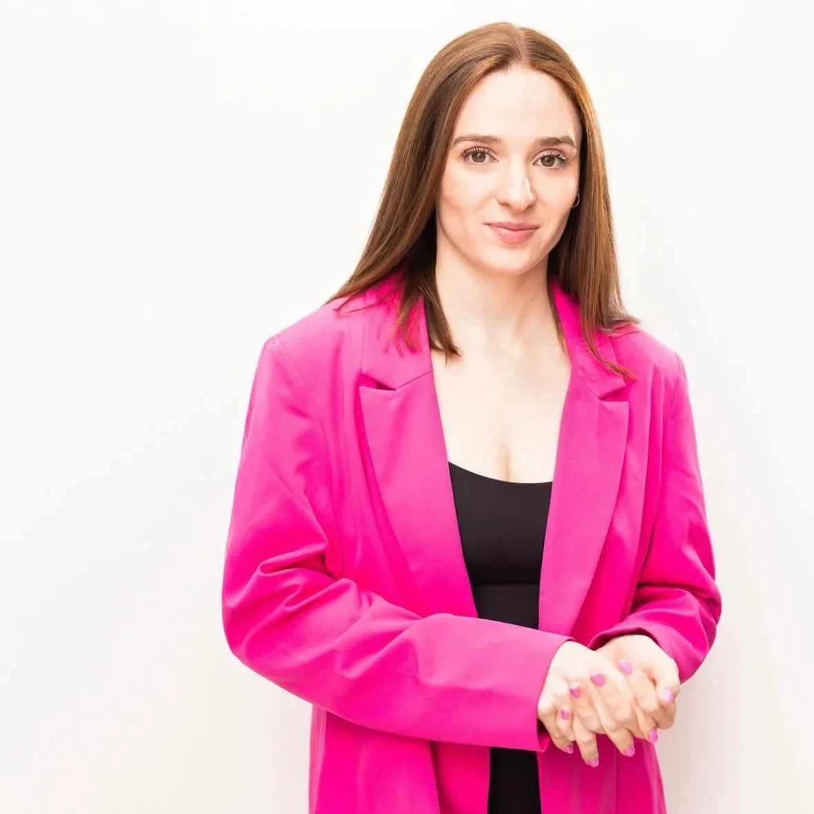 A woman with long brown hair wearing a pink blazer over a black top standing against a plain white background.