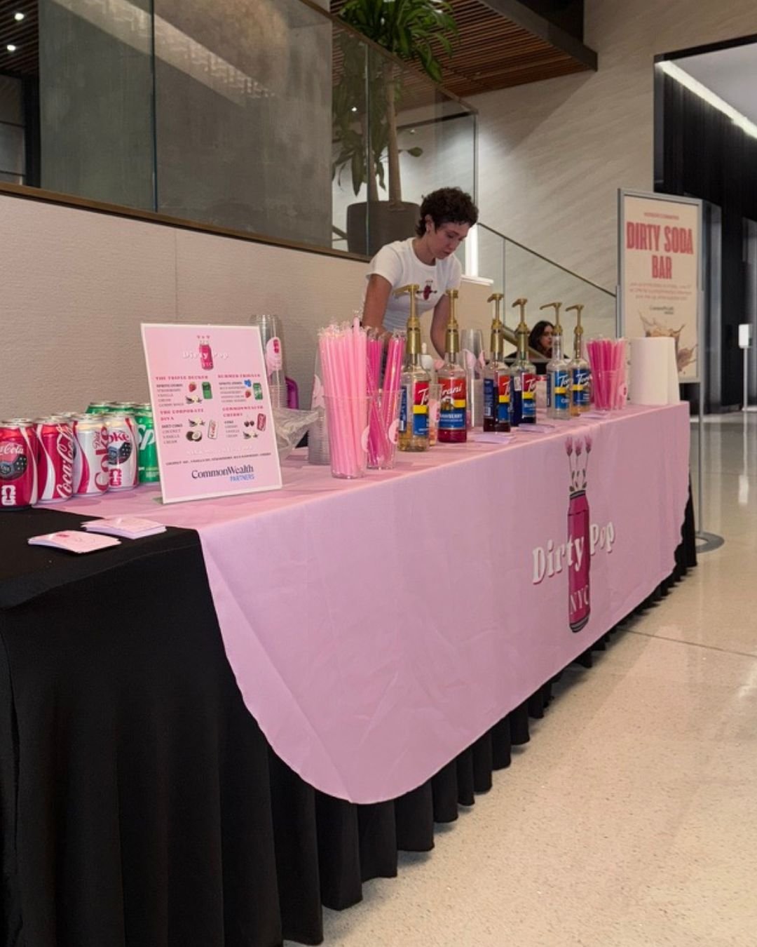 A pink tablecloth-covered stand serving drinks with a sign for Dirty Pop, featuring pink napkins, straws, and various bottled beverages, in an indoor setting with a woman preparing drinks behind the table.