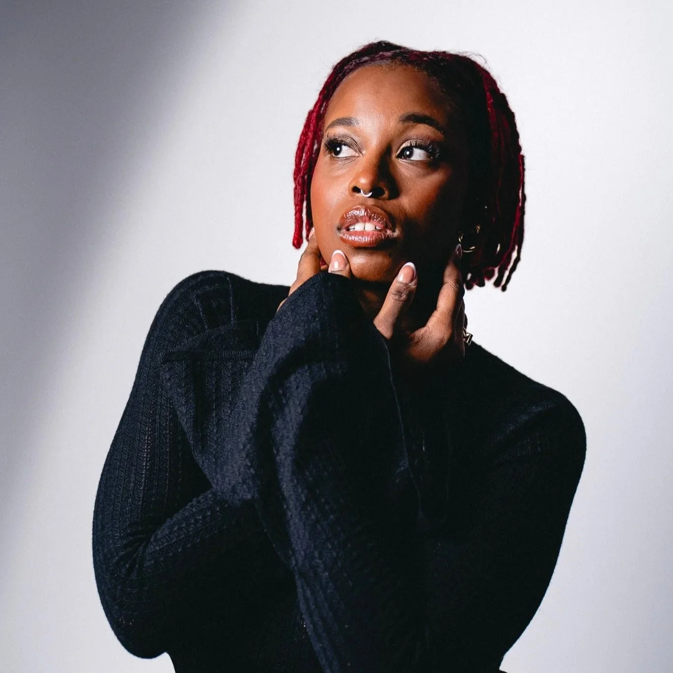 A woman with red dreadlocks poses against a plain background, wearing a black textured jacket, with one hand touching her neck and the other hand near her face.