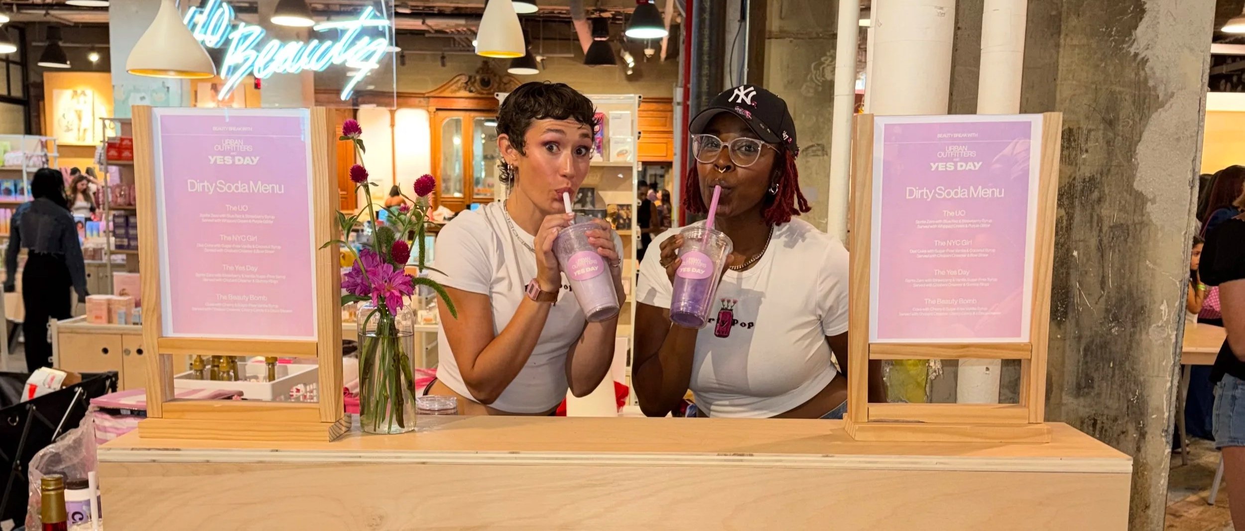 Two women standing behind a booth, drinking purple beverages with straws, in a store or market setting.