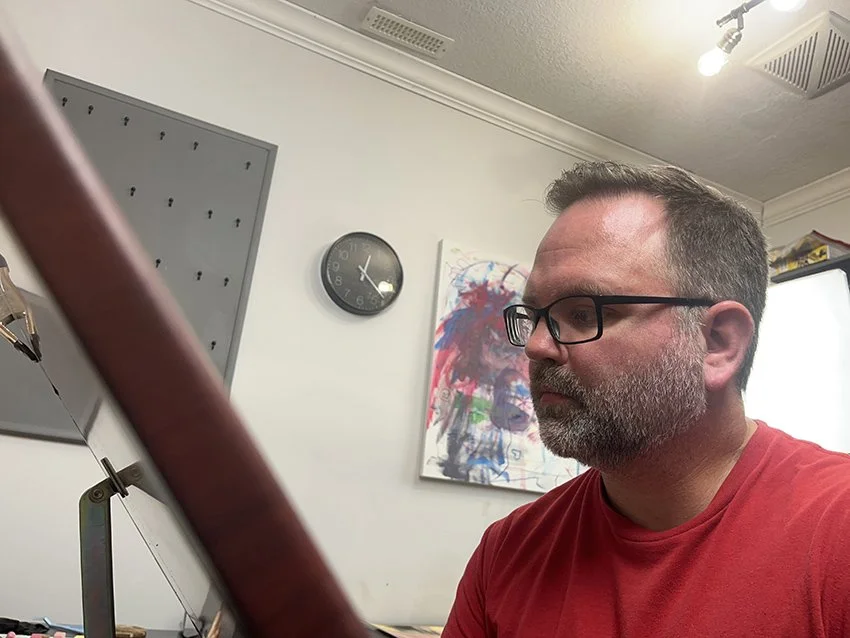 A man with glasses, a beard, and short hair working at a desk in an office, with a clock and a colorful abstract painting on the wall behind him.