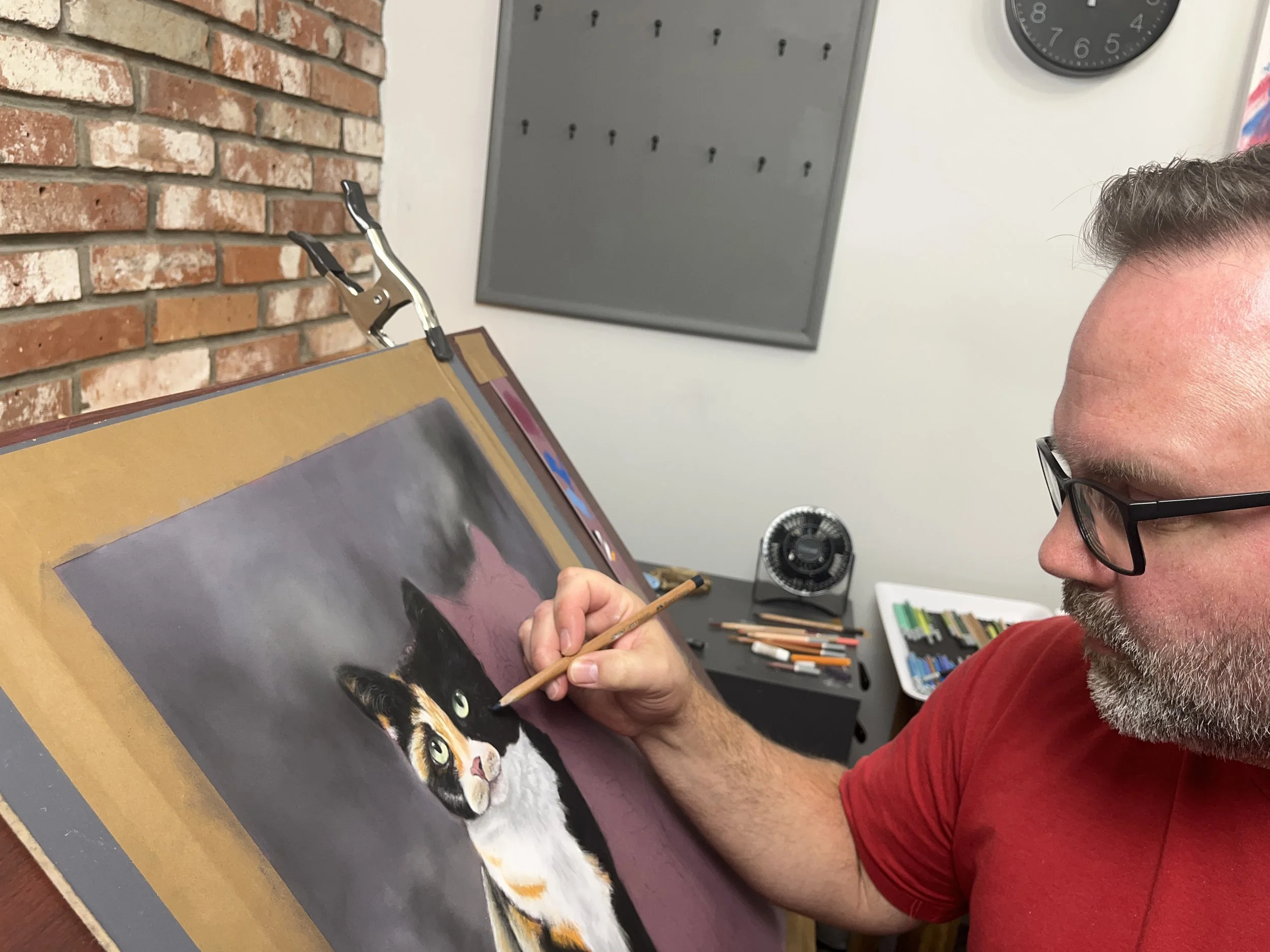 A man with glasses and a beard, wearing a red shirt, is painting a realistic portrait of a cat on canvas.