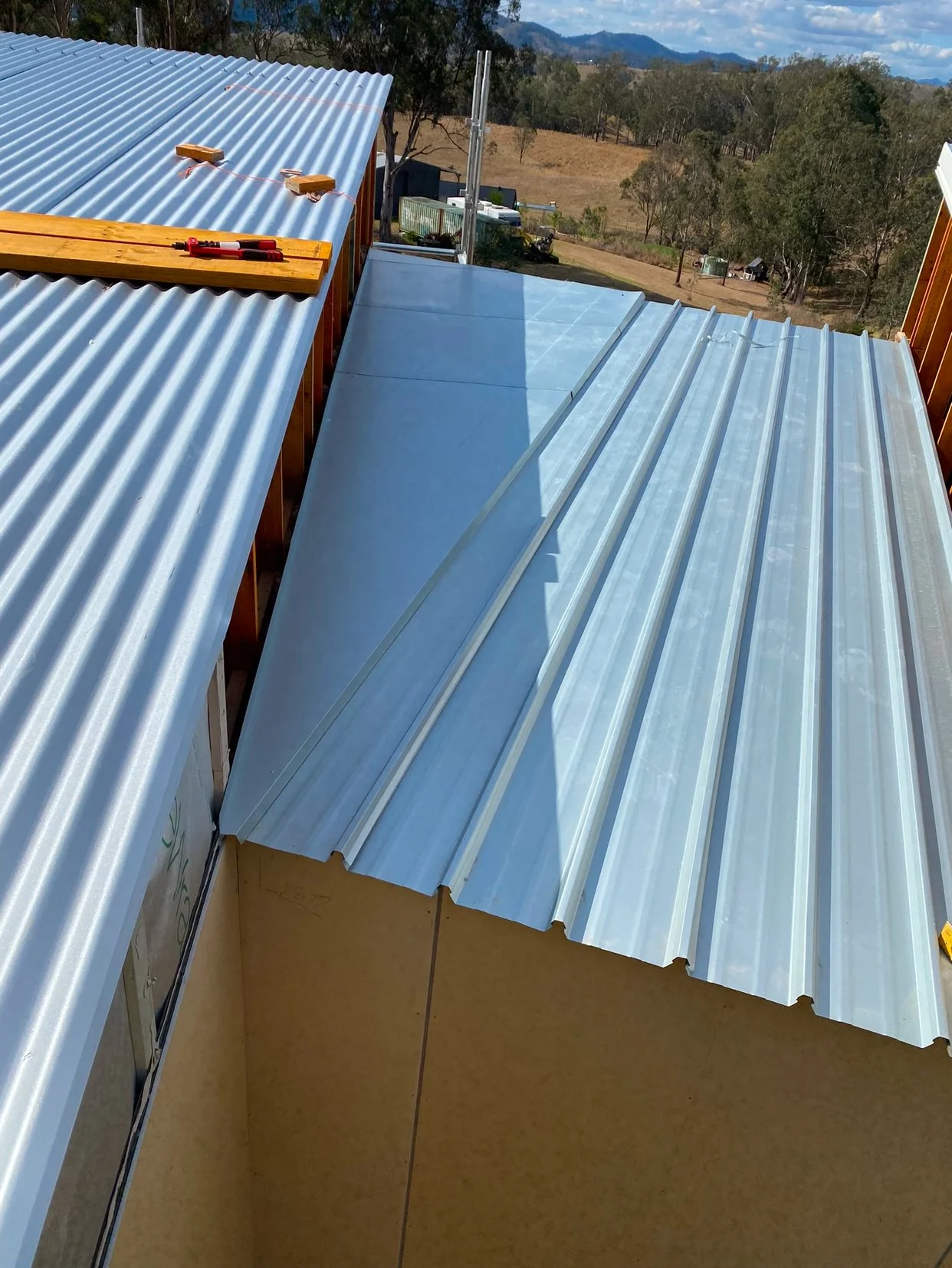 Construction site showing metal roofing sheets being installed on a building with views of trees, hills, and mountains in the background.