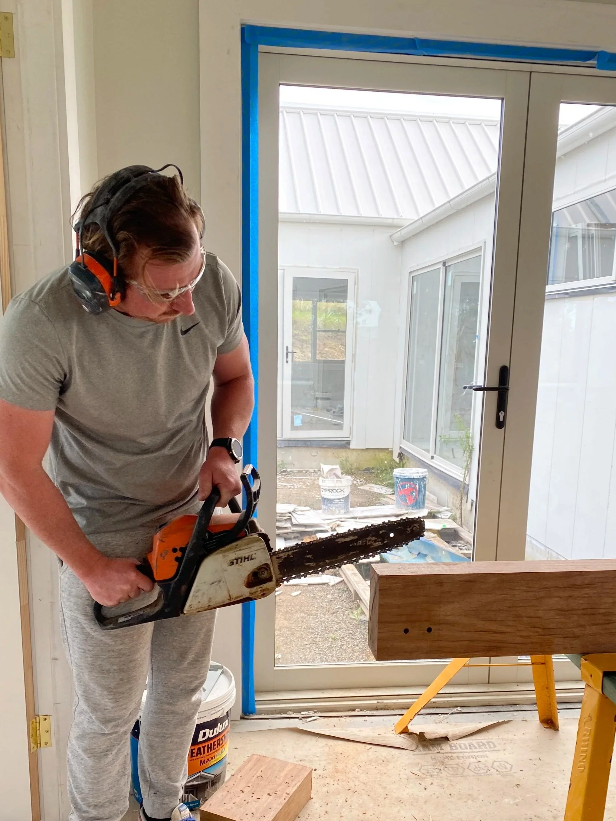 A person wearing safety glasses, ear protection, and a gray Nike t-shirt, using a chainsaw to cut wood inside a room under construction, with construction materials and tools around.