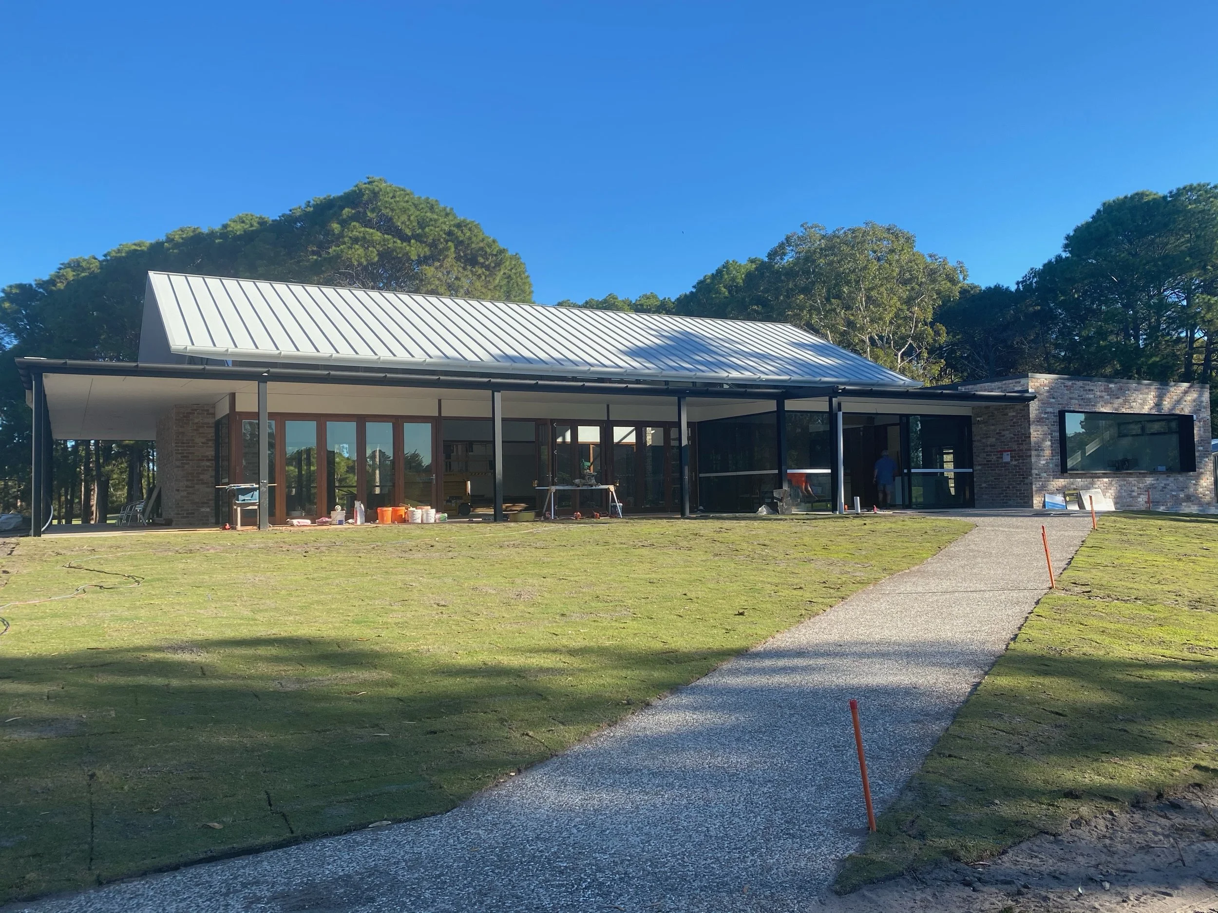 Construction site with a modern house featuring large glass windows, brick accents, a metal roof, and a covered patio, surrounded by green grass and trees, with a bright blue sky.
