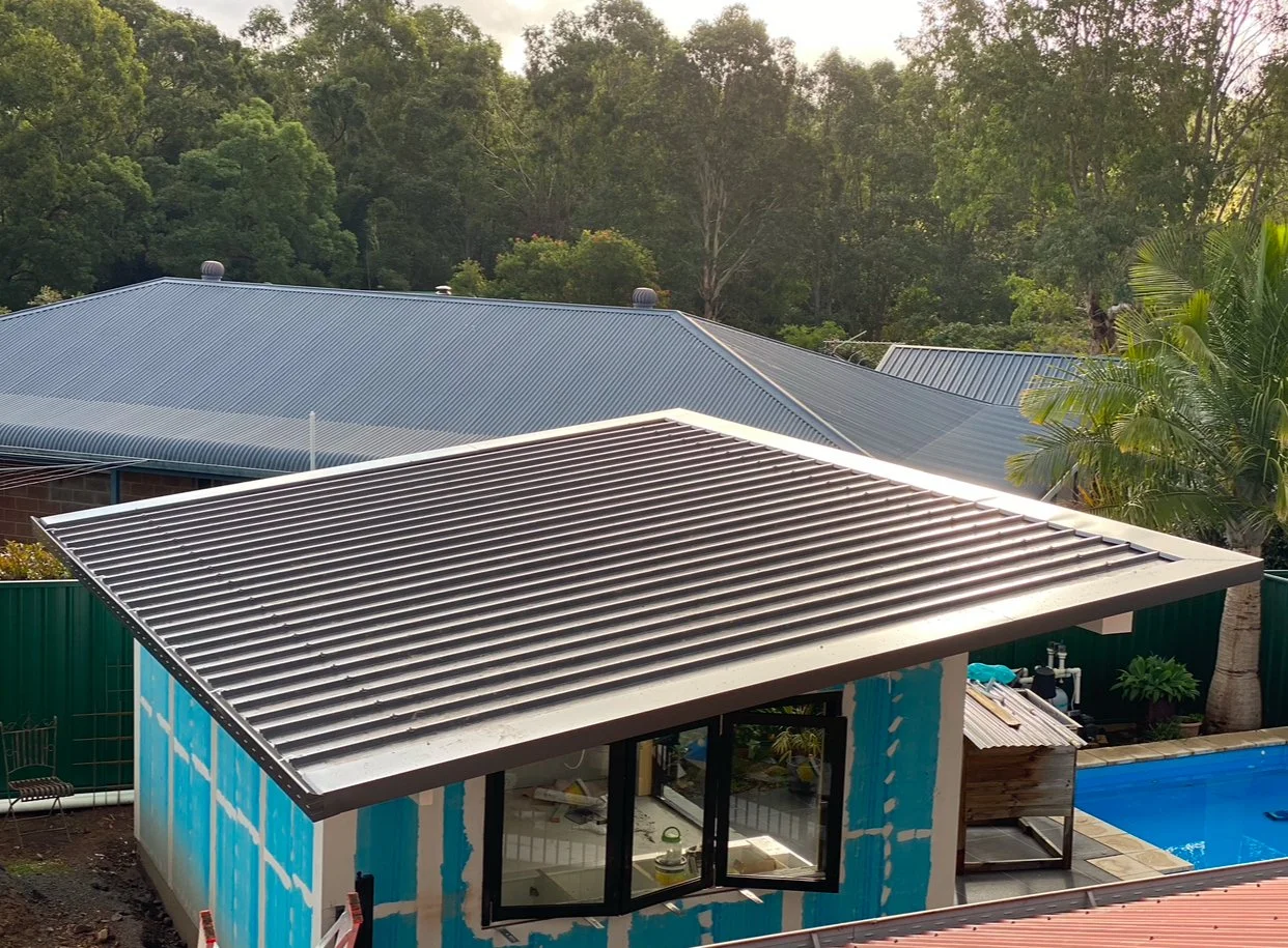 Construction of a small building with a metal roof beside a swimming pool, surrounded by lush trees and green fencing.