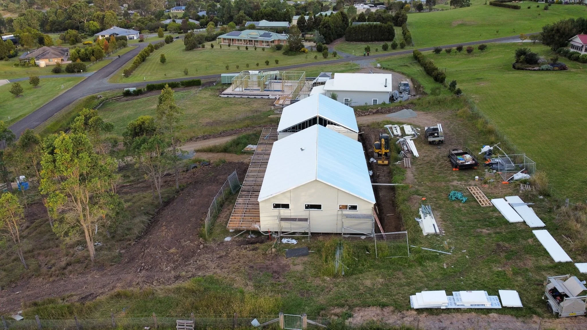 Aerial view of a residential construction site with two new houses being built, surrounded by green lawns and trees, and neighboring houses in the background.