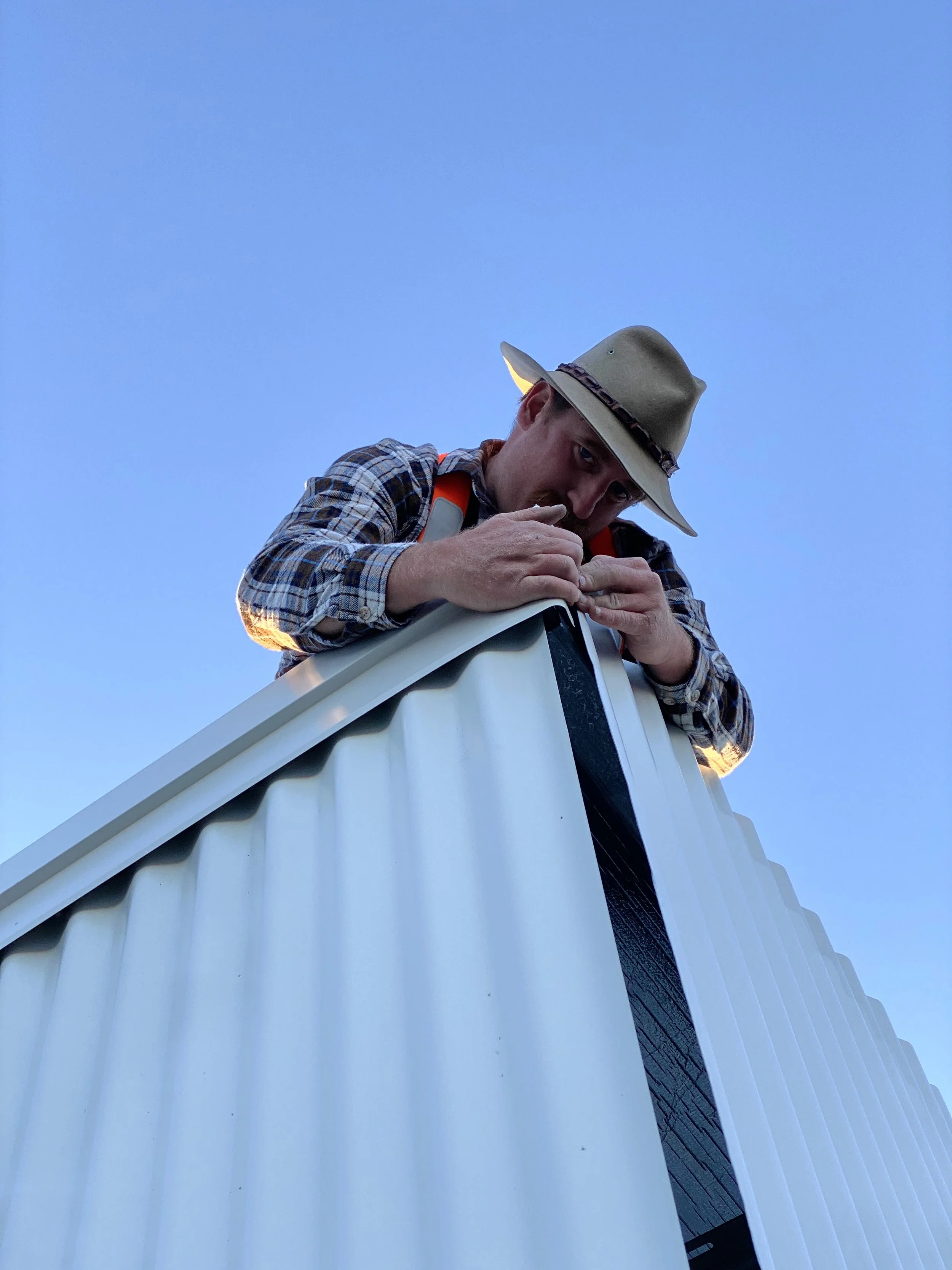 A man wearing a wide-brimmed hat and plaid shirt leaning over a white structure, against a clear blue sky.