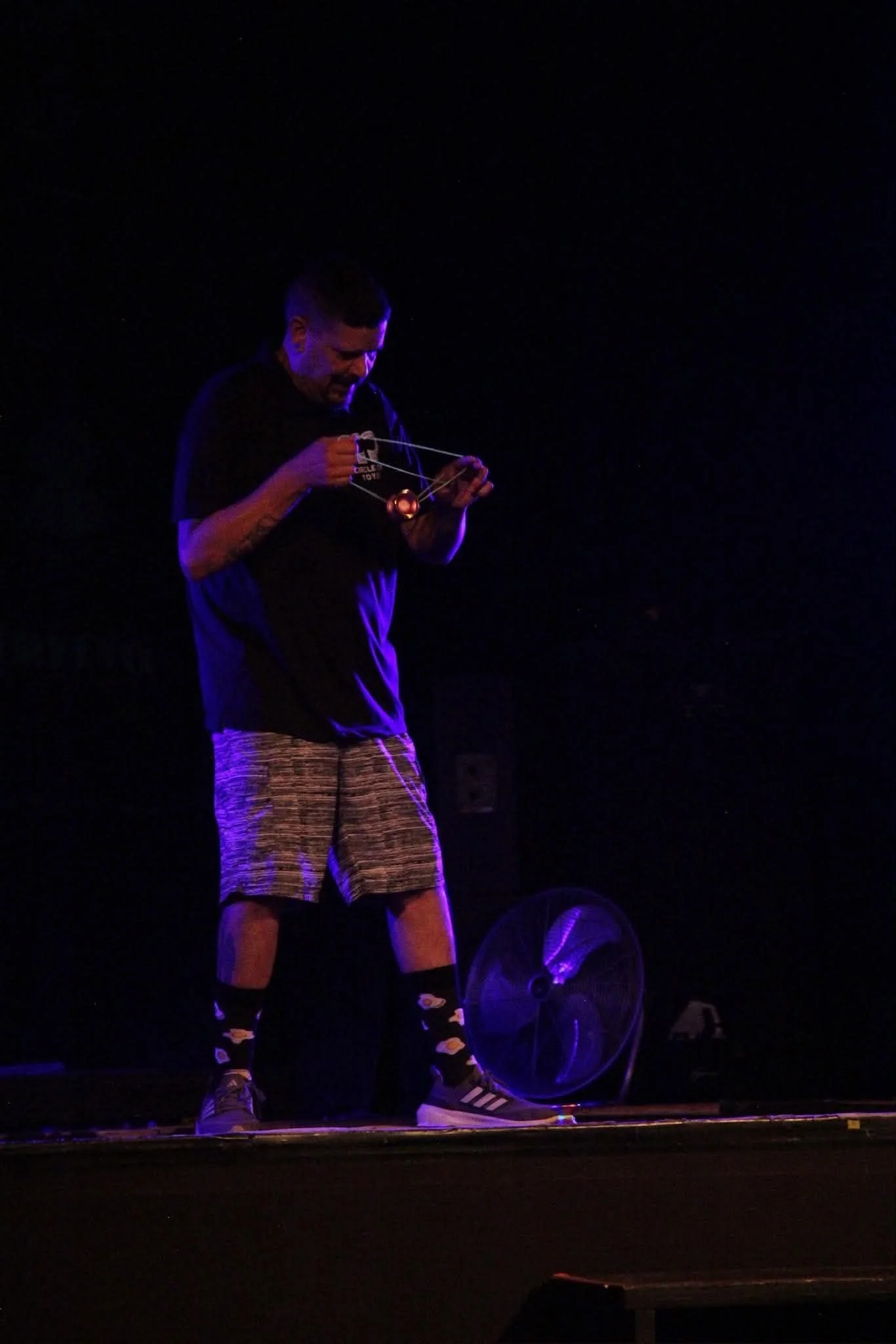 Performer in shorts and sneakers manipulating a glowing yo-yo on a dark stage under purple lighting