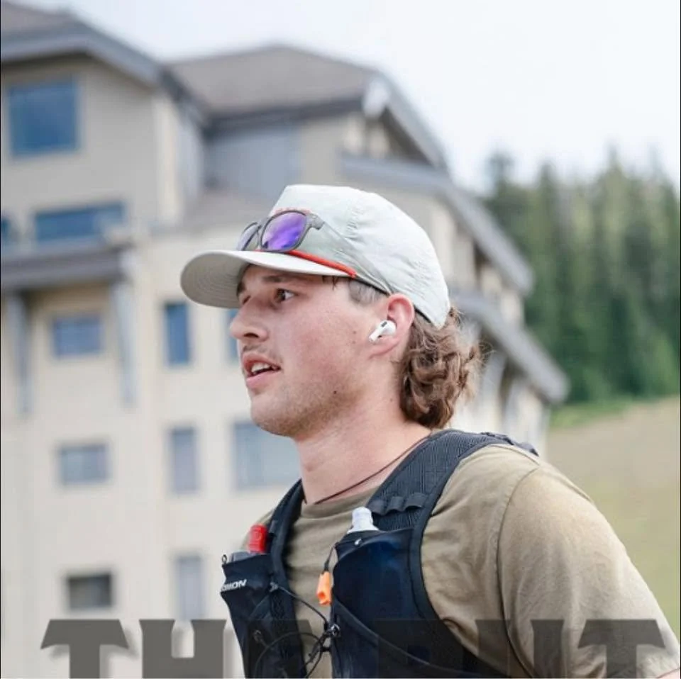 Trail runner wearing a cap, sunglasses, earbuds, and hydration vest outdoors