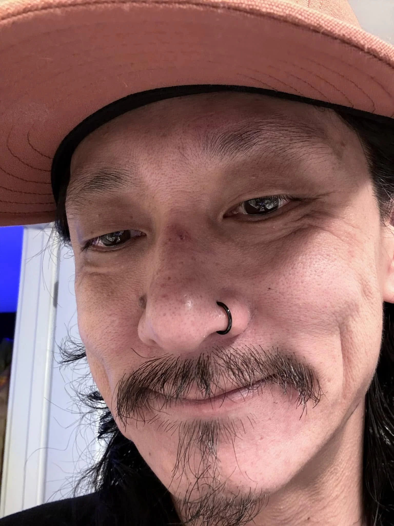 Close-up portrait of a person wearing a pink hat with a black nose ring and mustache