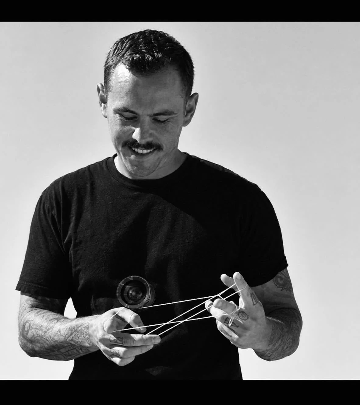 Black-and-white portrait of a tattooed man in a black T-shirt manipulating white strings attached to a small camera lens.