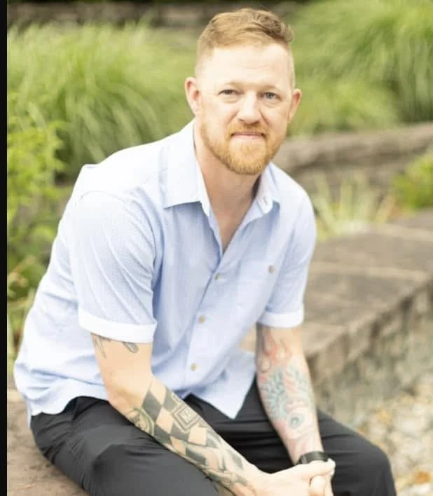 Bearded man with tattooed arms sitting outdoors in a light blue shirt and black pants