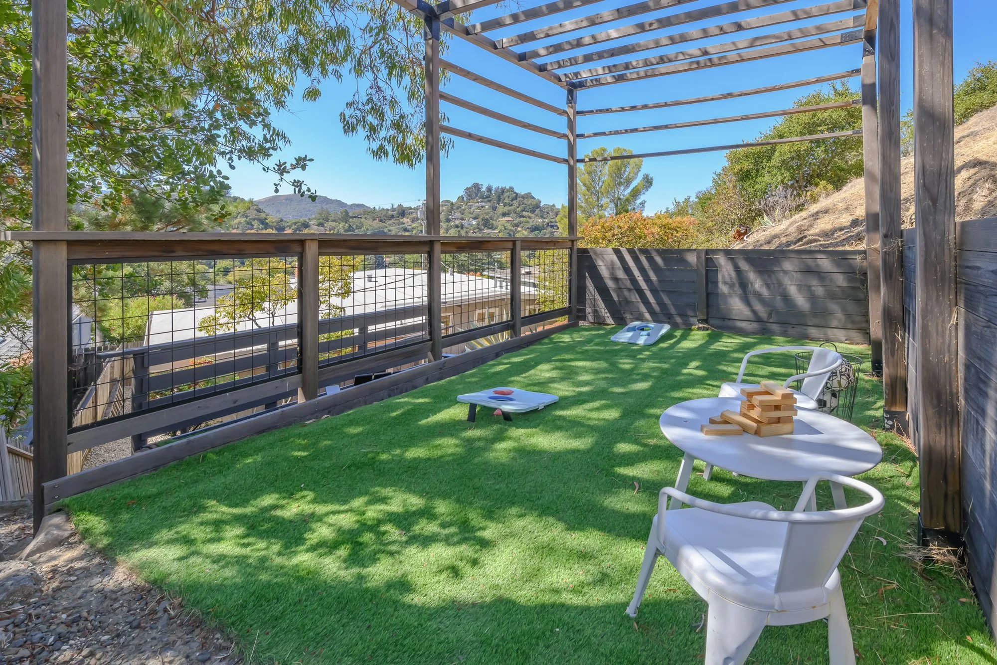 A backyard patio with artificial grass, a few white chairs, a small table with wooden blocks, and green trees with hills in the distance under a clear blue sky.