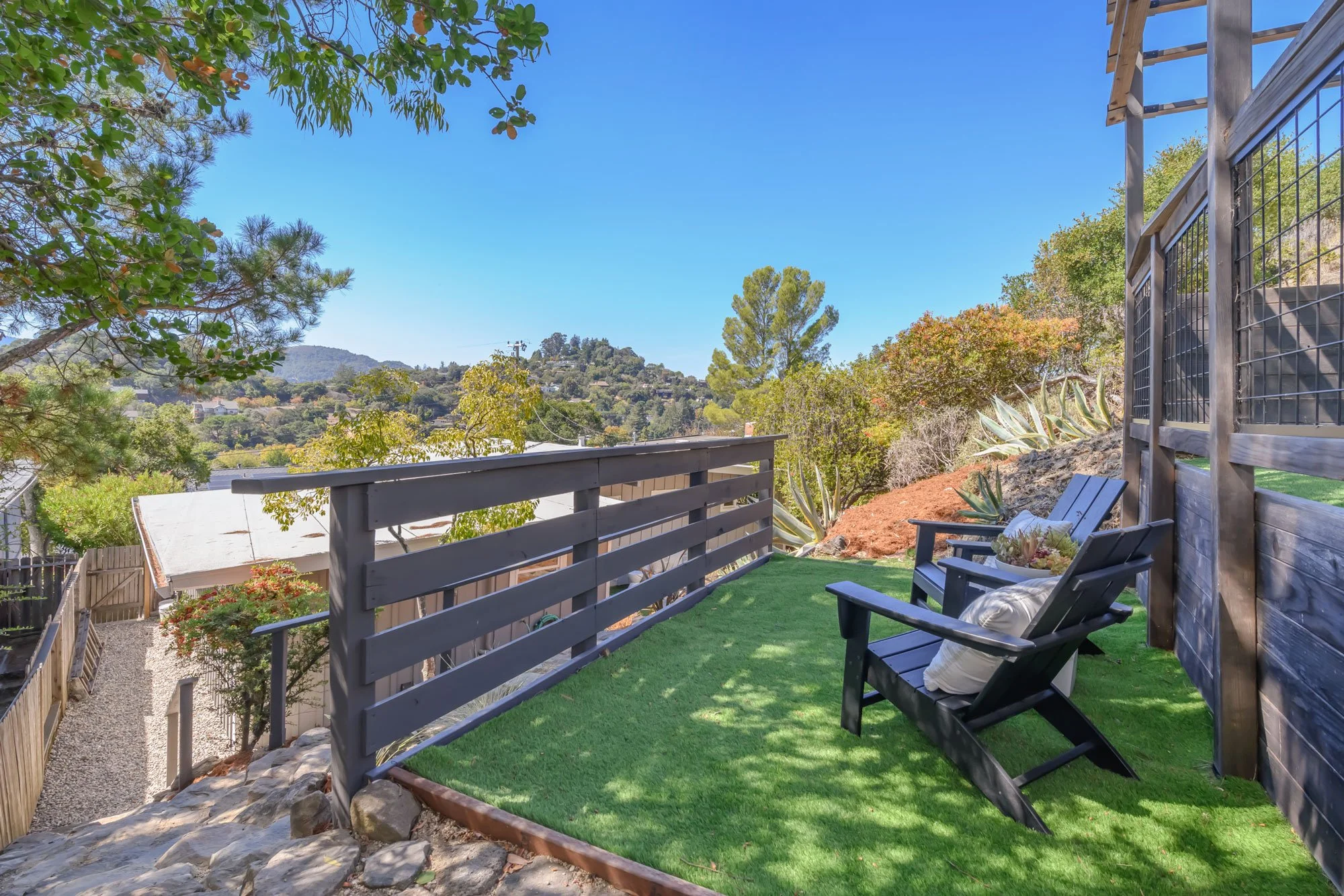 A backyard patio with artificial grass and outdoor chairs, overlooking a hillside with trees and houses under a clear blue sky.